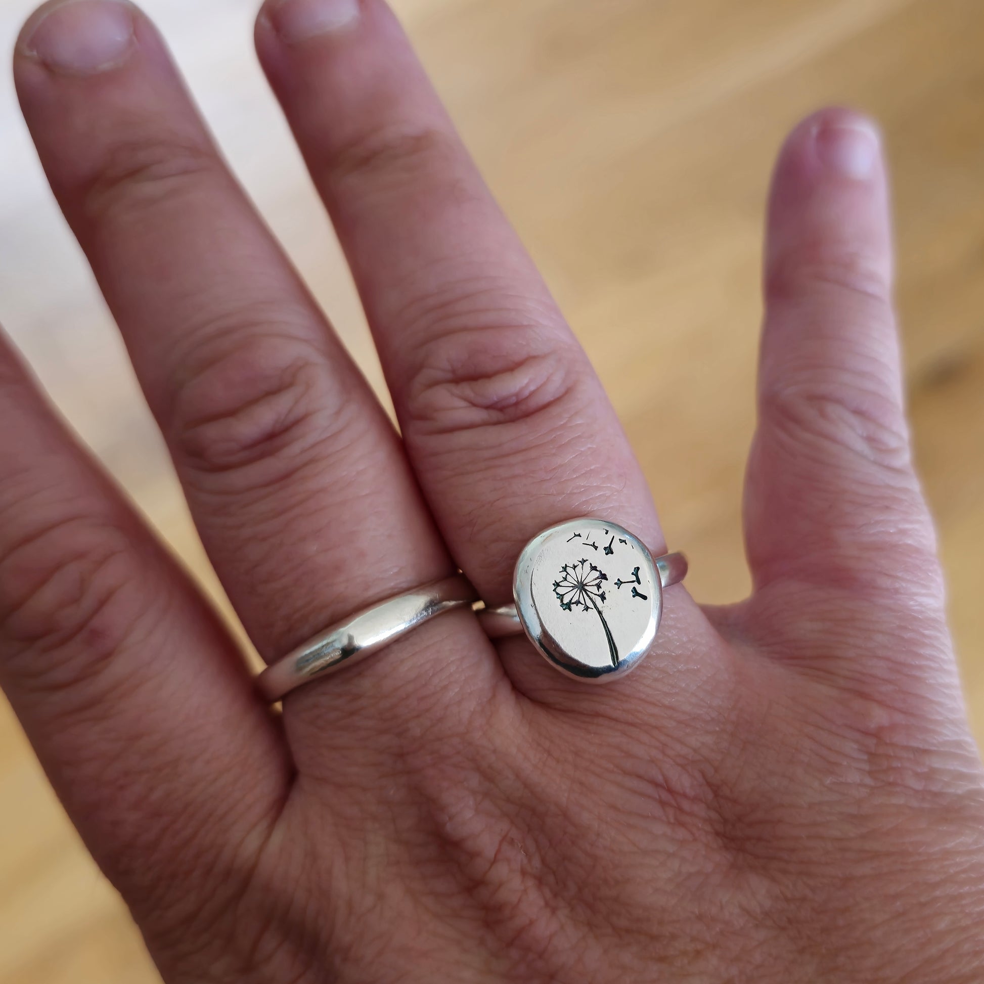 A silver ring with a plain band and a flat pebble top with a dandelion clock and fluff engraved on it. Shown on a hand.