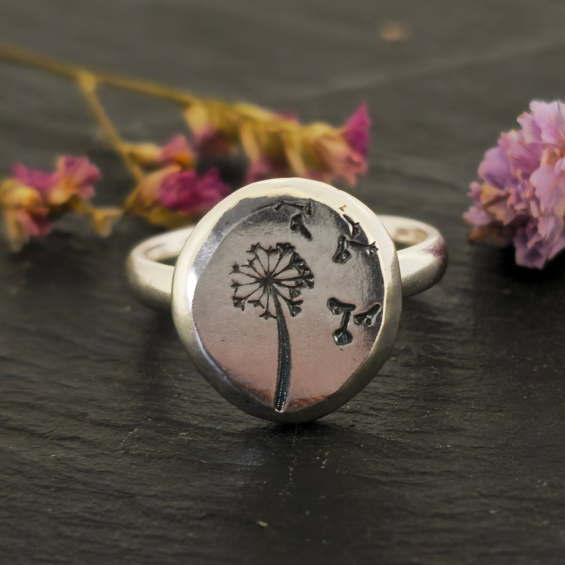 A silver ring with a plain band and a flat pebble top with a dandelion clock and fluff engraved on it. Shown on slate with flowers.