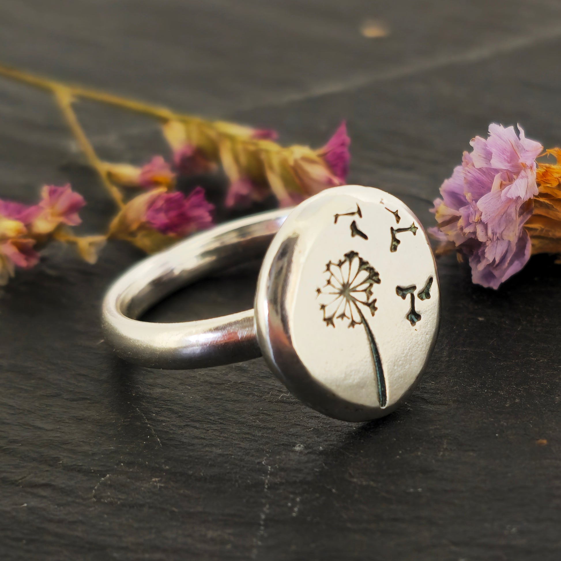 A silver ring with a plain band and a flat pebble top with a dandelion clock and fluff engraved on it. Shown on slate with flowers.