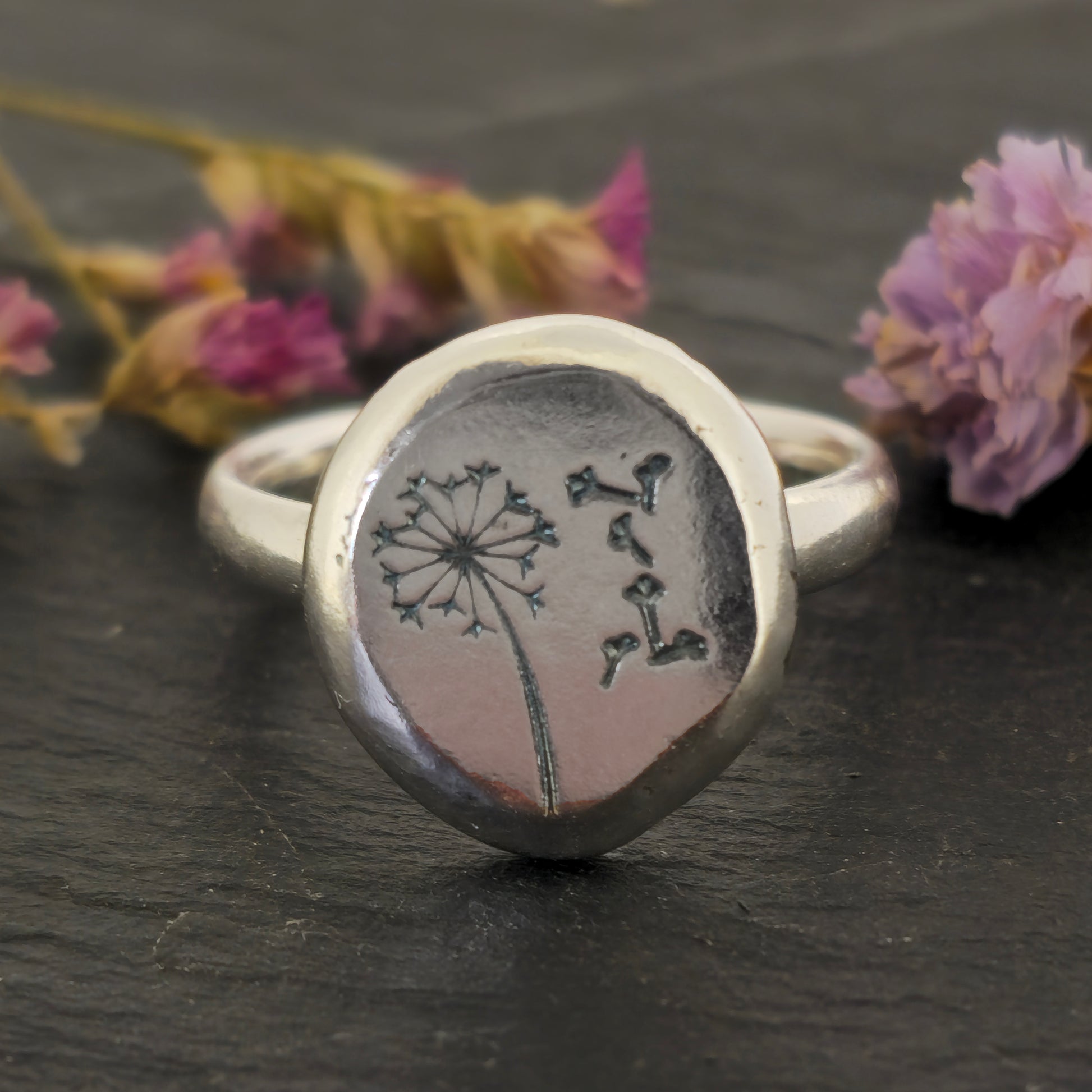 A silver ring with a plain band and a flat pebble top with a dandelion clock and fluff engraved on it. Shown on slate with flowers.