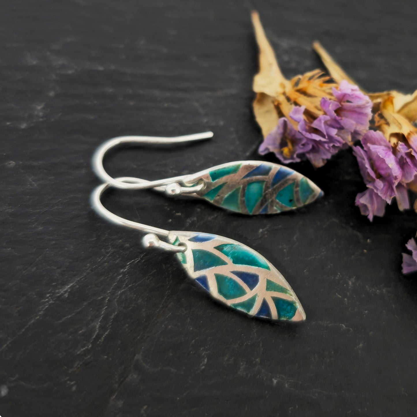 Silver drop earrings. Each earrings has a marquise shape with geometric cells containing blue, green and turquoise enamel. Pictured on slate with flowers.