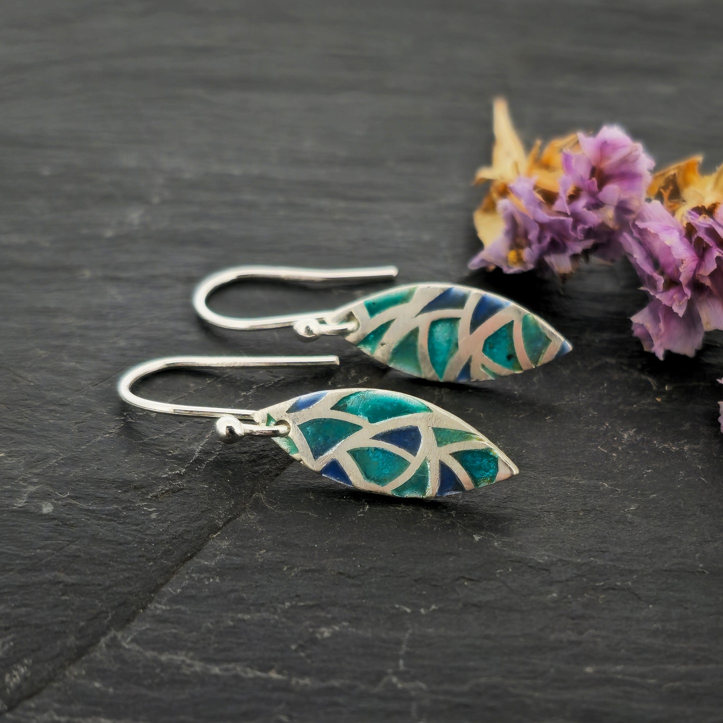Silver drop earrings. Each earrings has a marquise shape with geometric cells containing blue, green and turquoise enamel. Pictured on slate with flowers.