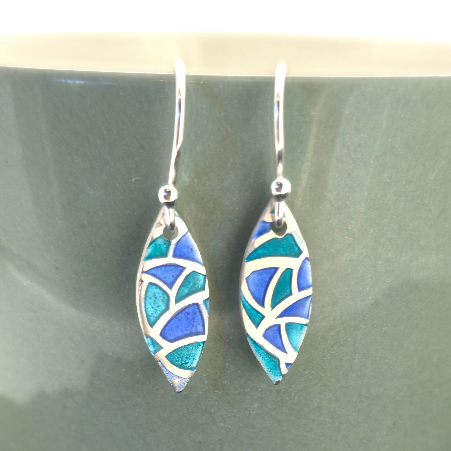Silver drop earrings. Each earrings has a marquise shape with geometric cells containing blue, green and turquoise enamel. Pictured on a green bowl.