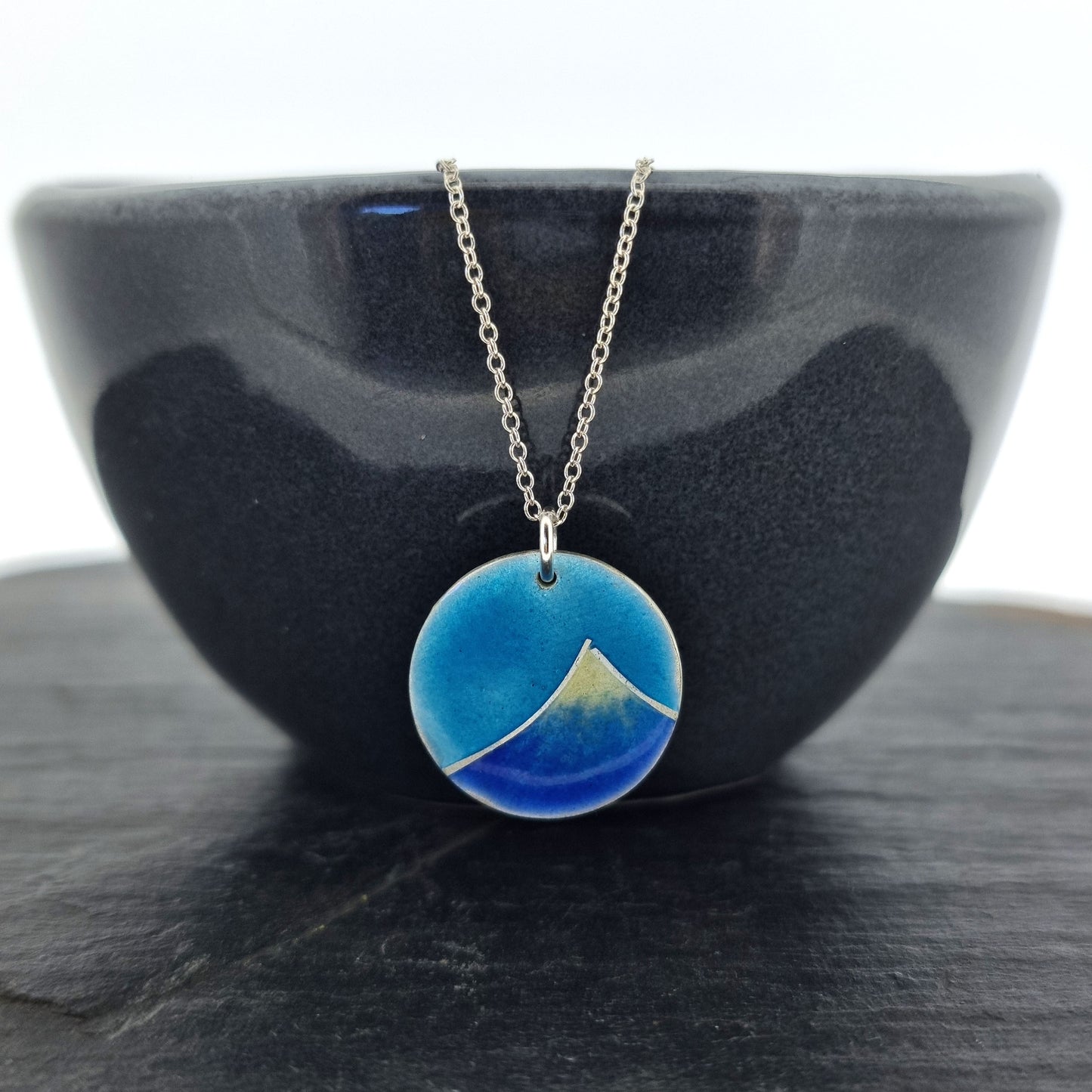 Silver and enamel round pendant featuring a dark blue and white mountain with a turquoise blue sky. On a silver chain. Pictured on a bowl.