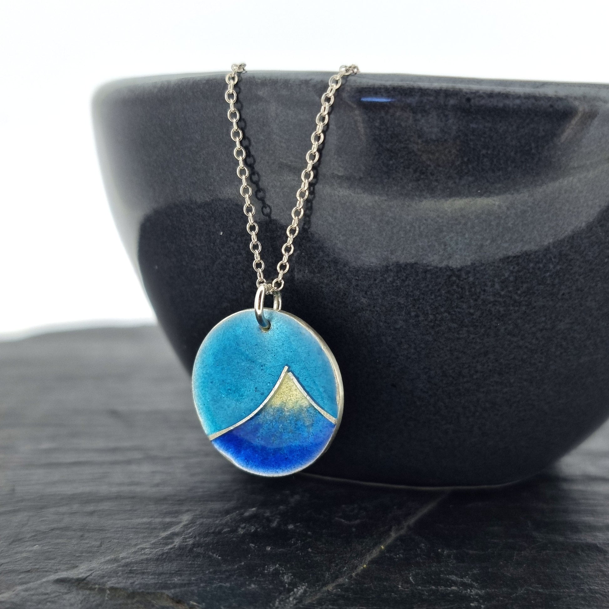 Silver and enamel round pendant featuring a dark blue and white mountain with a turquoise blue sky. On a silver chain. Pictured on a bowl.