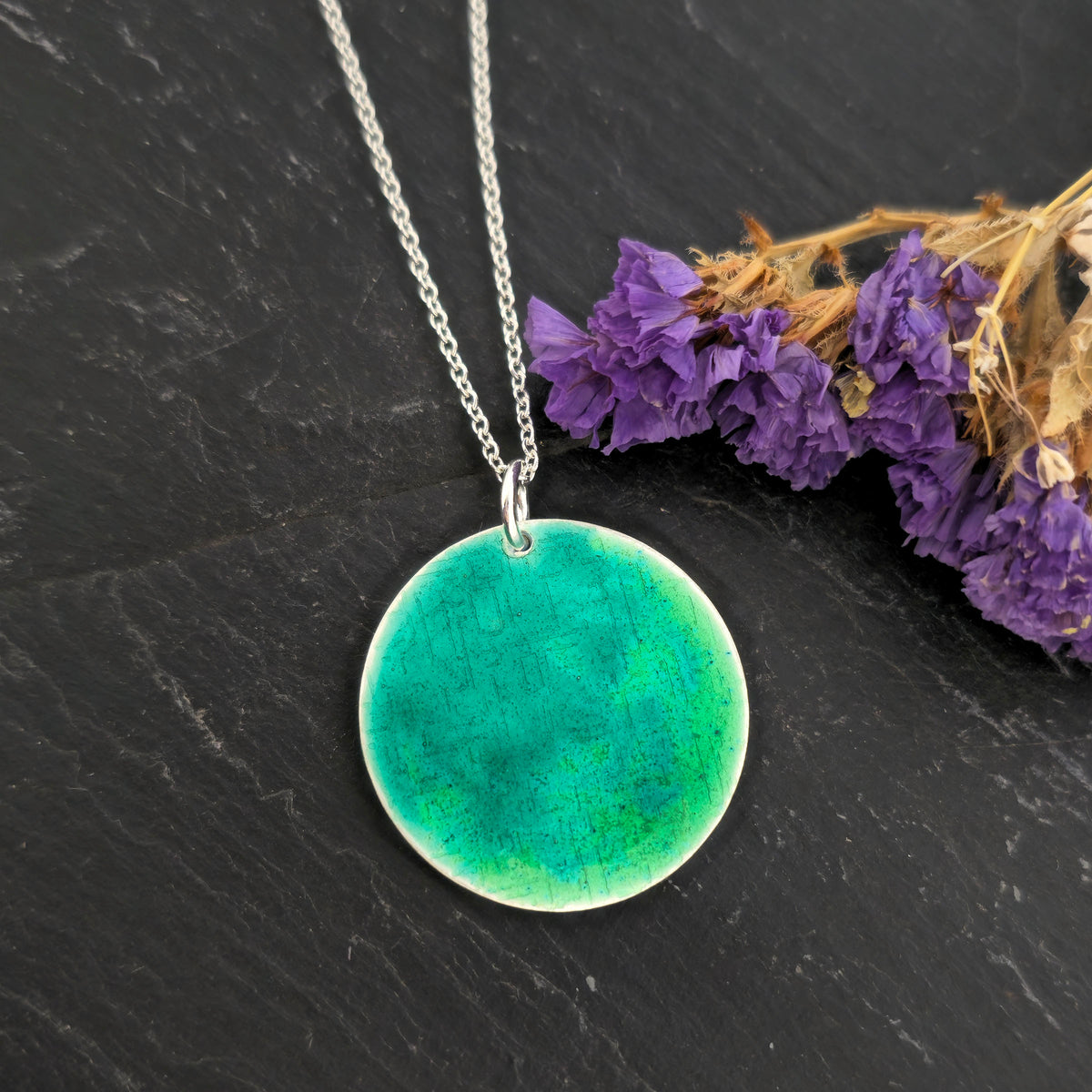 A silver chain necklace with a handmade round silver disc pendant featuring a vibrant green and turquoise enamelled textured pattern, displayed on a dark slate surface. Mijoux Jewellery.