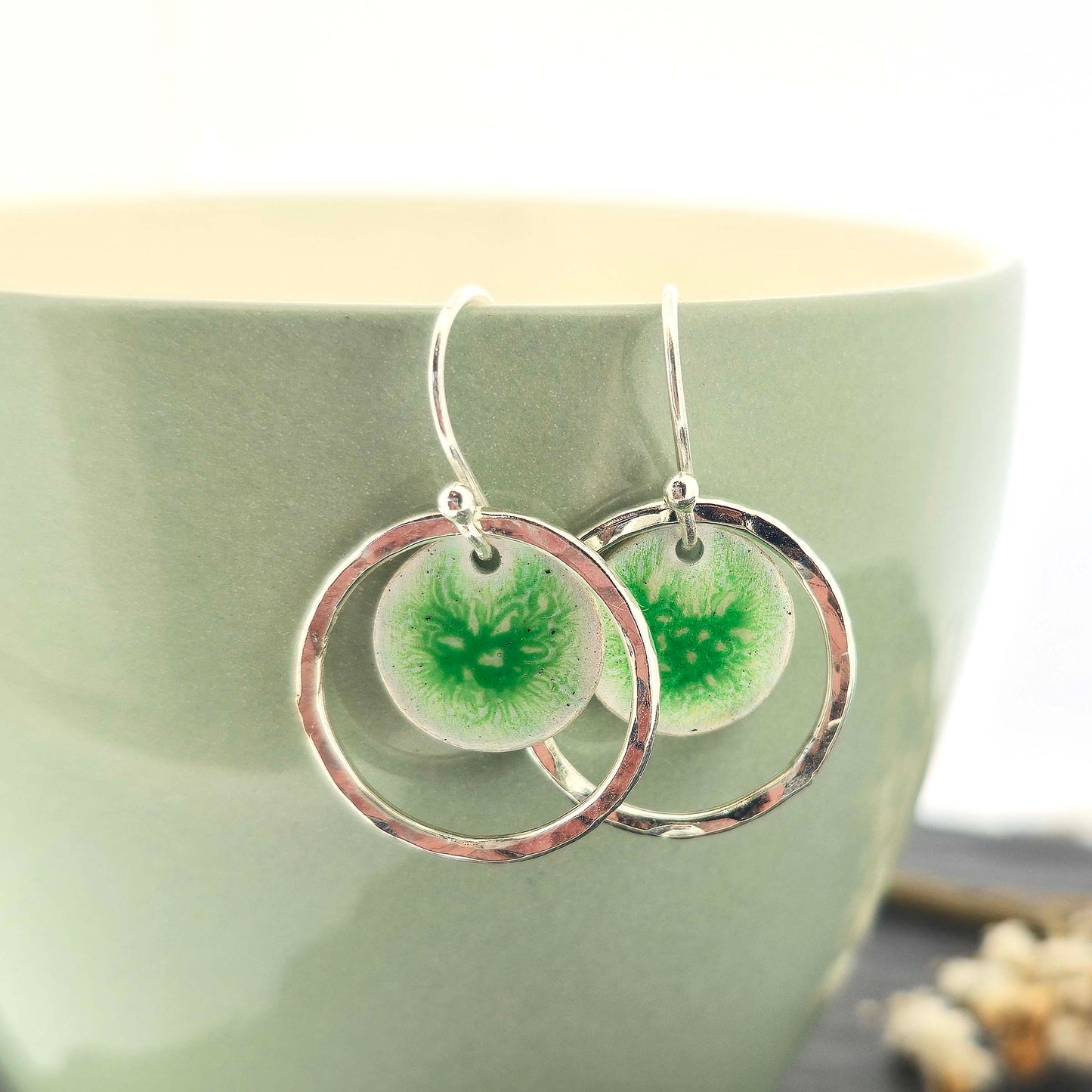 Silver drop earrings with a central green patterned enamel disc and a surrounding hammered silver circle. Shown on a green cup.