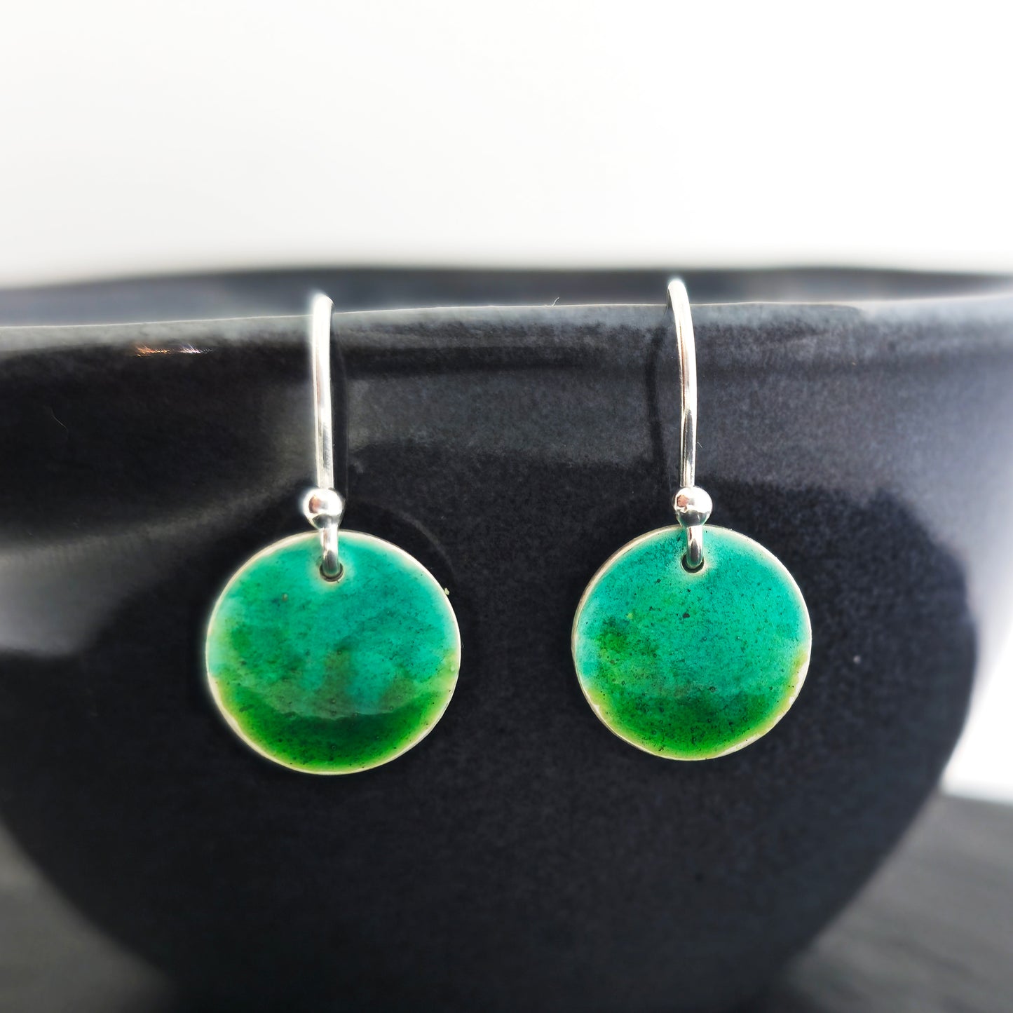 Round silver drop earrings with a mix of green enamels. Pictured on a black bowl.