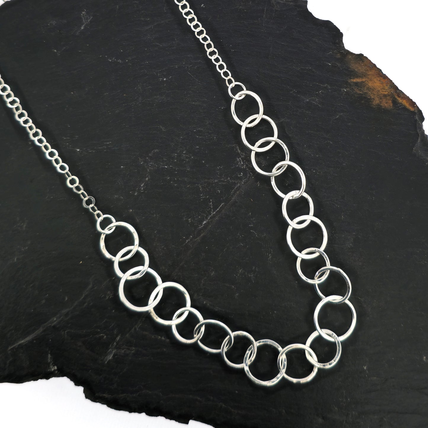 A silver necklace made up of different size circles with a hammered finish and smaller silver chain at the back. Pictured on slate.