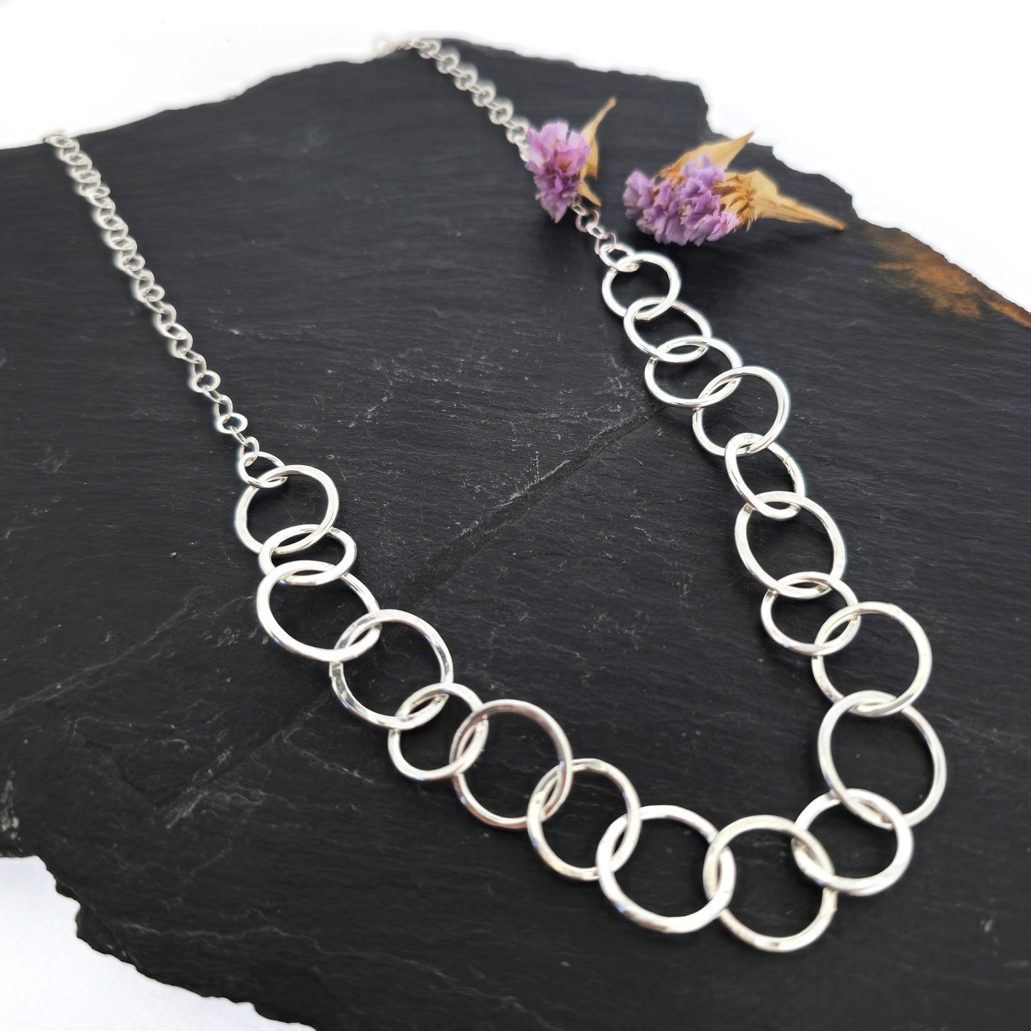 A silver necklace made up of different size circles with a hammered finish and smaller silver chain at the back. Pictured on slate with flowers.