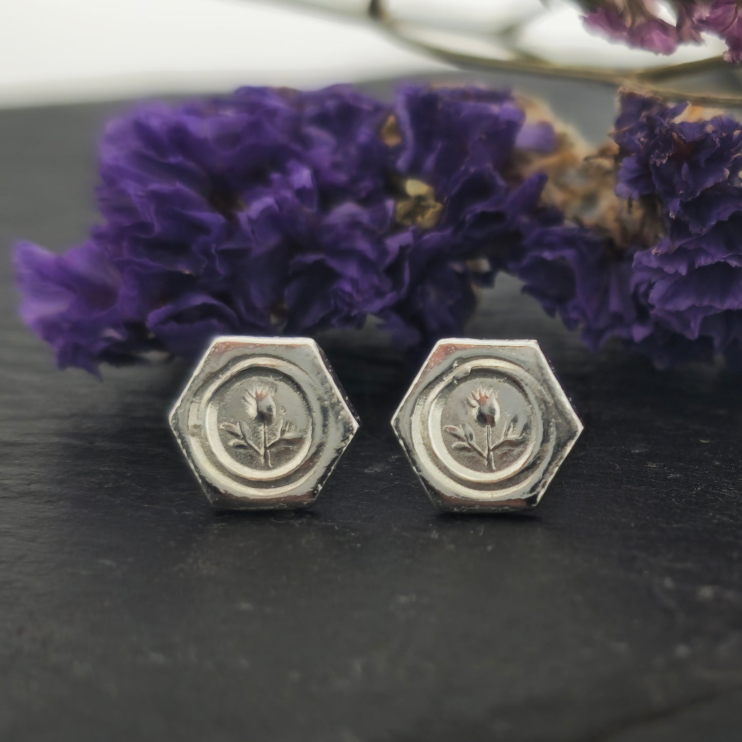 A pair of hexagon shaped sterling silver stud earrings with a thistle on them. Shown on slate with flowers.