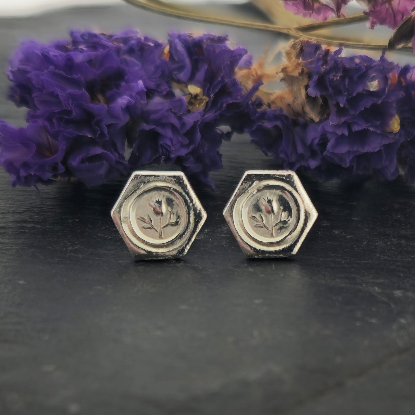 A pair of hexagon shaped sterling silver stud earrings with a thistle on them. Shown on slate with flowers.
