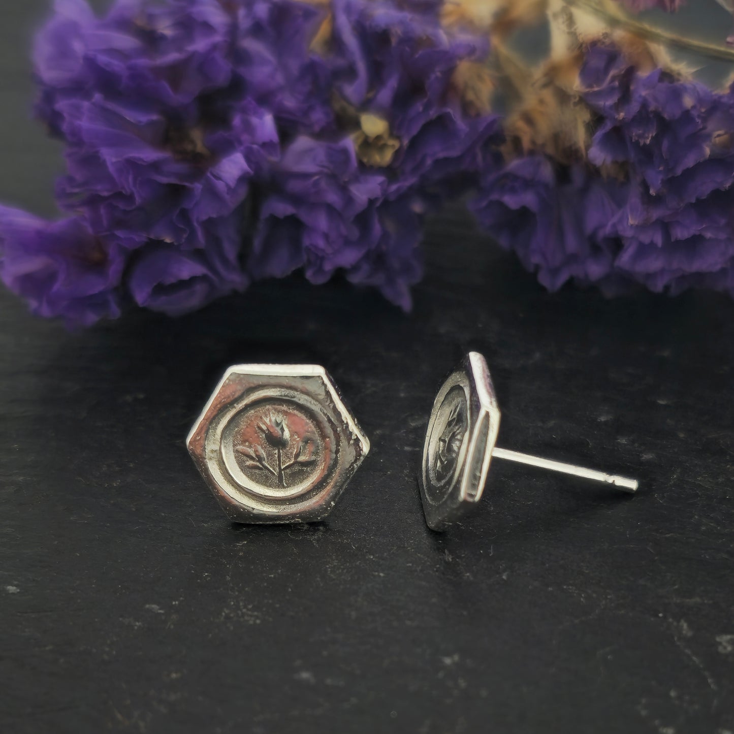 A pair of hexagon shaped sterling silver stud earrings with a thistle on them. Shown on slate with flowers.