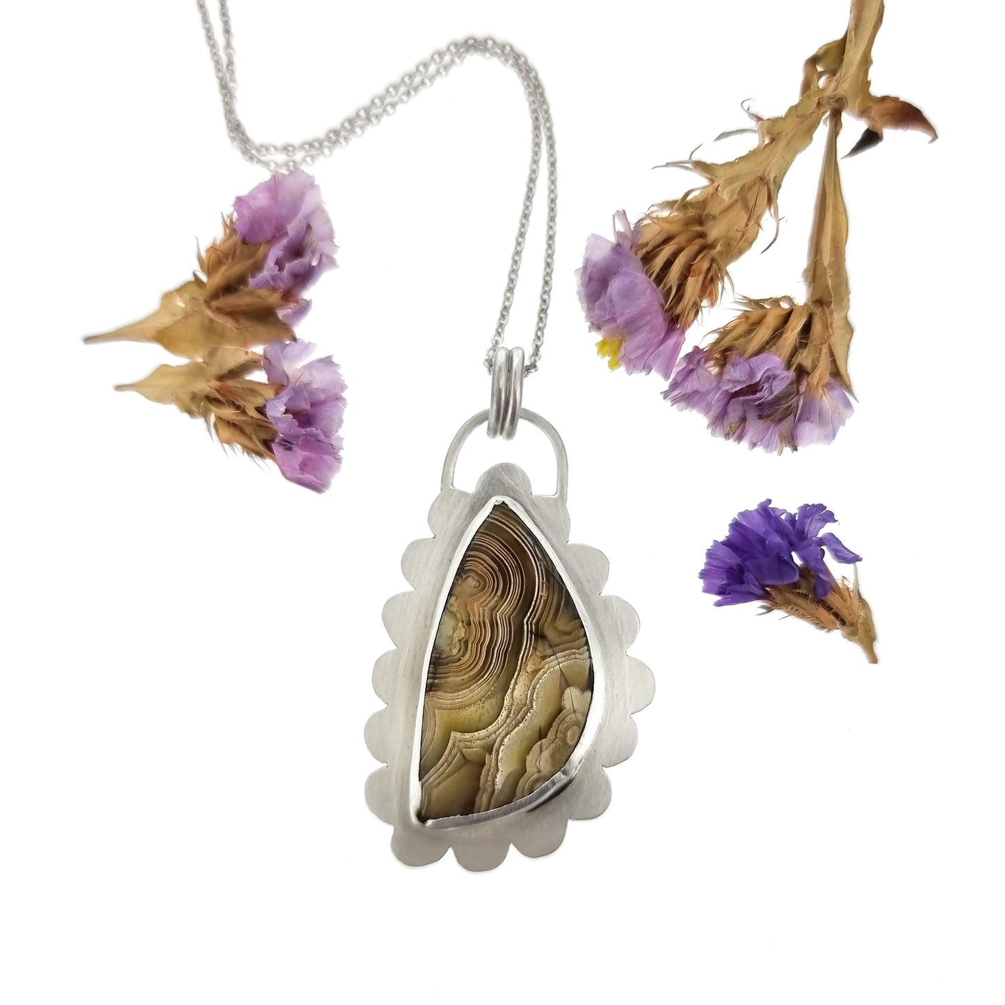 Silver pendant with brown jasper gemstone with curbed striations in a silver surround shaped like a cloud. On a silver chain. Pictured with flowers.