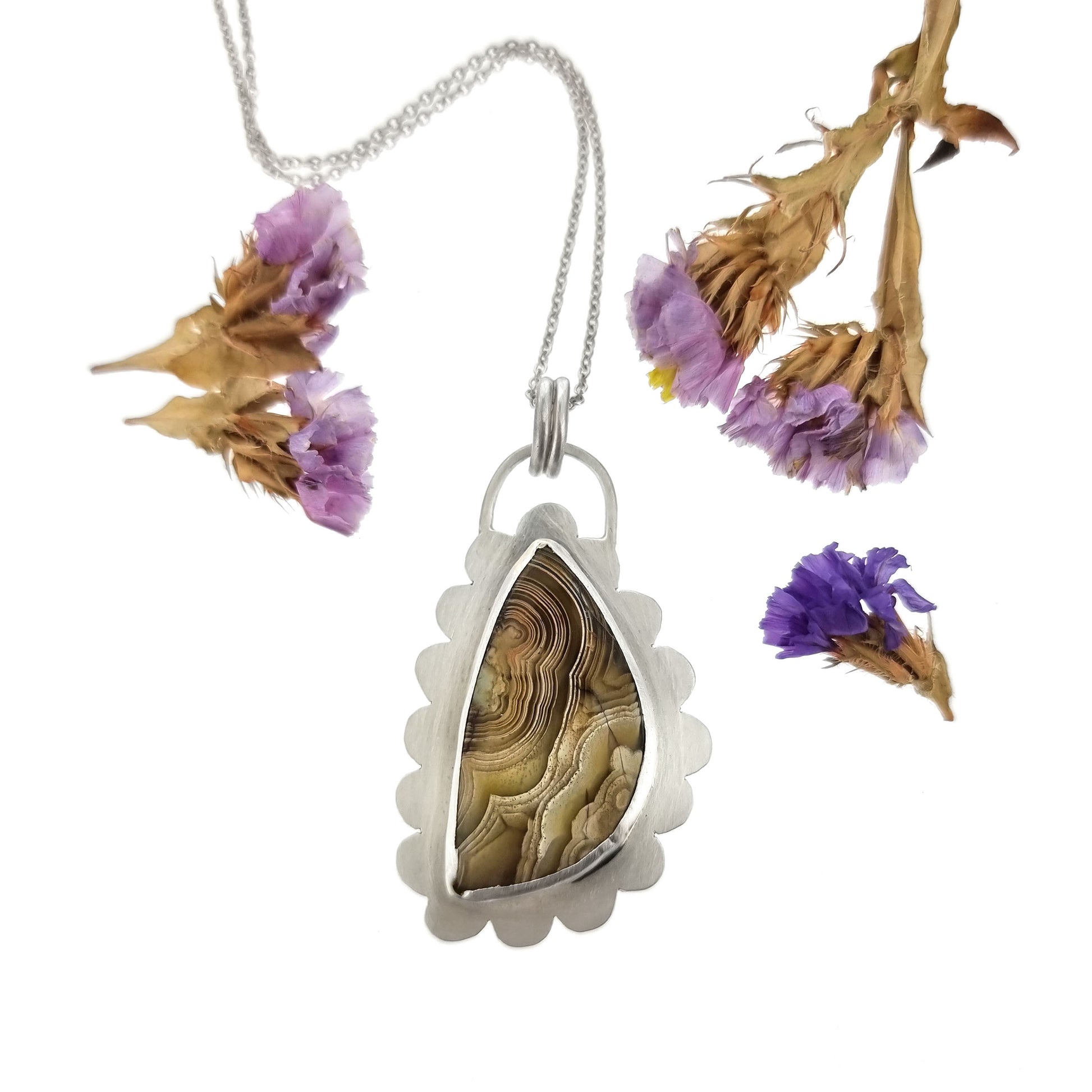 Silver pendant with brown jasper gemstone with curbed striations in a silver surround shaped like a cloud. On a silver chain. Pictured with flowers.
