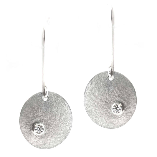 Silver round drop earrings with a matte texture and a sparkly clear CZ set off centre. On long ear hooks. Front view.