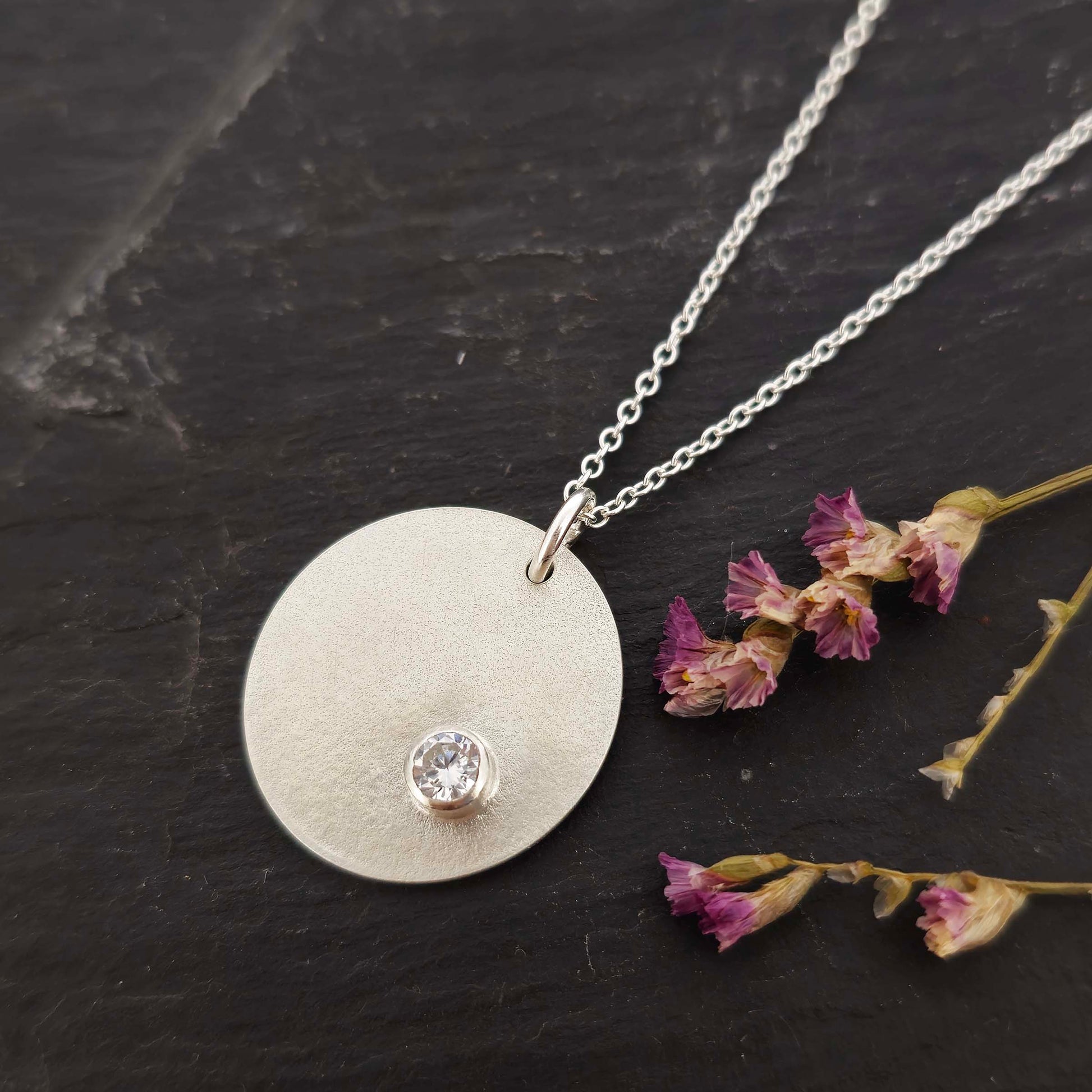 A silver round disc pendant with a matte finish and a sparkly clear CZ set off centre. On a silver chain. Pictured on slate with flowers.
