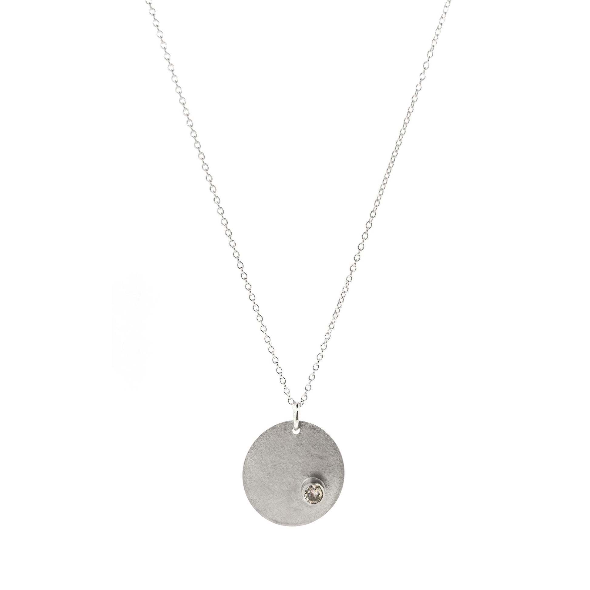 A silver round disc pendant with a matte finish and a sparkly clear CZ set off centre. On a silver chain. Front view.