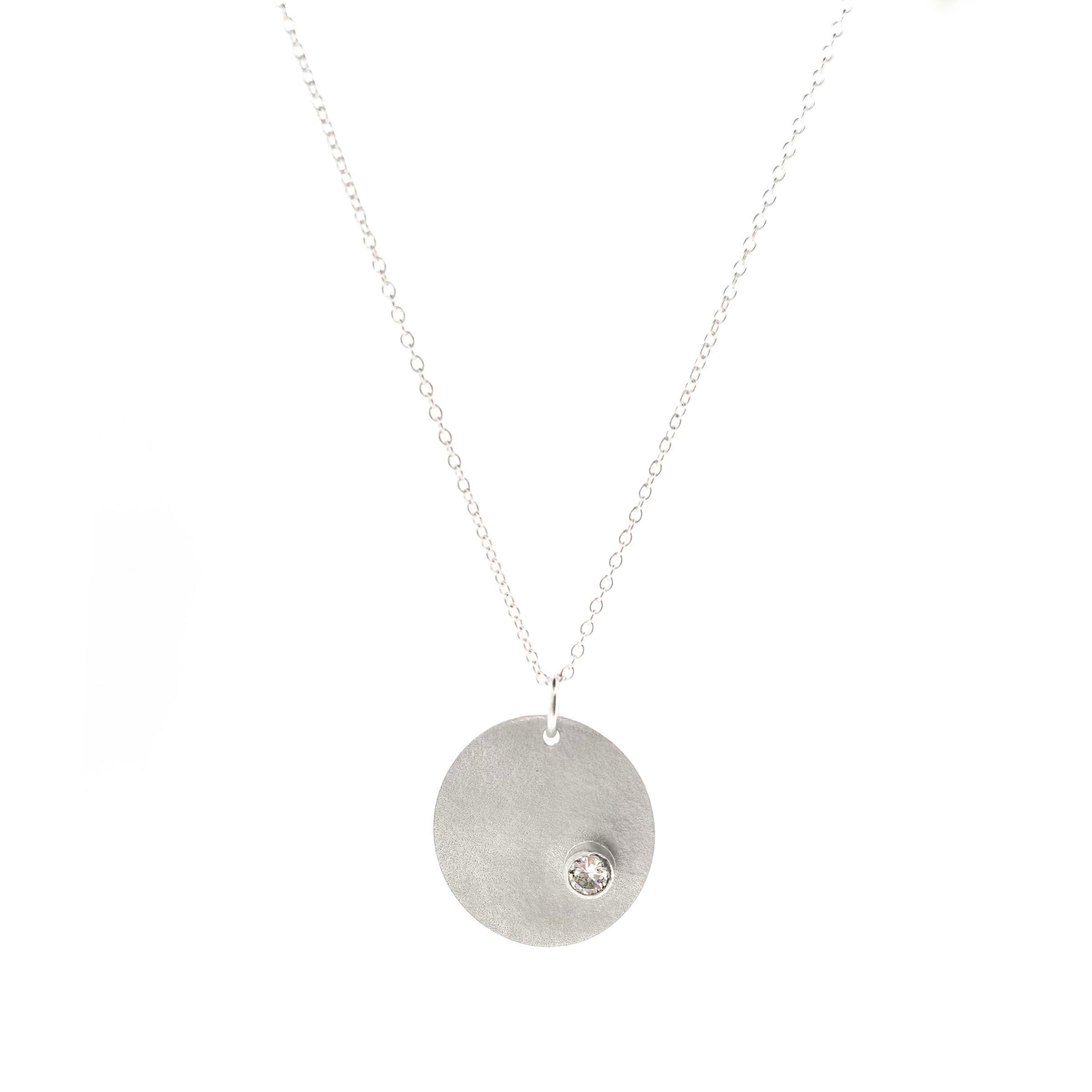 A silver round disc pendant with a matte finish and a sparkly clear CZ set off centre. On a silver chain. Front view.