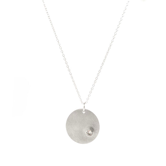 A silver round disc pendant with a matte finish and a sparkly clear CZ set off centre. On a silver chain. Front view.