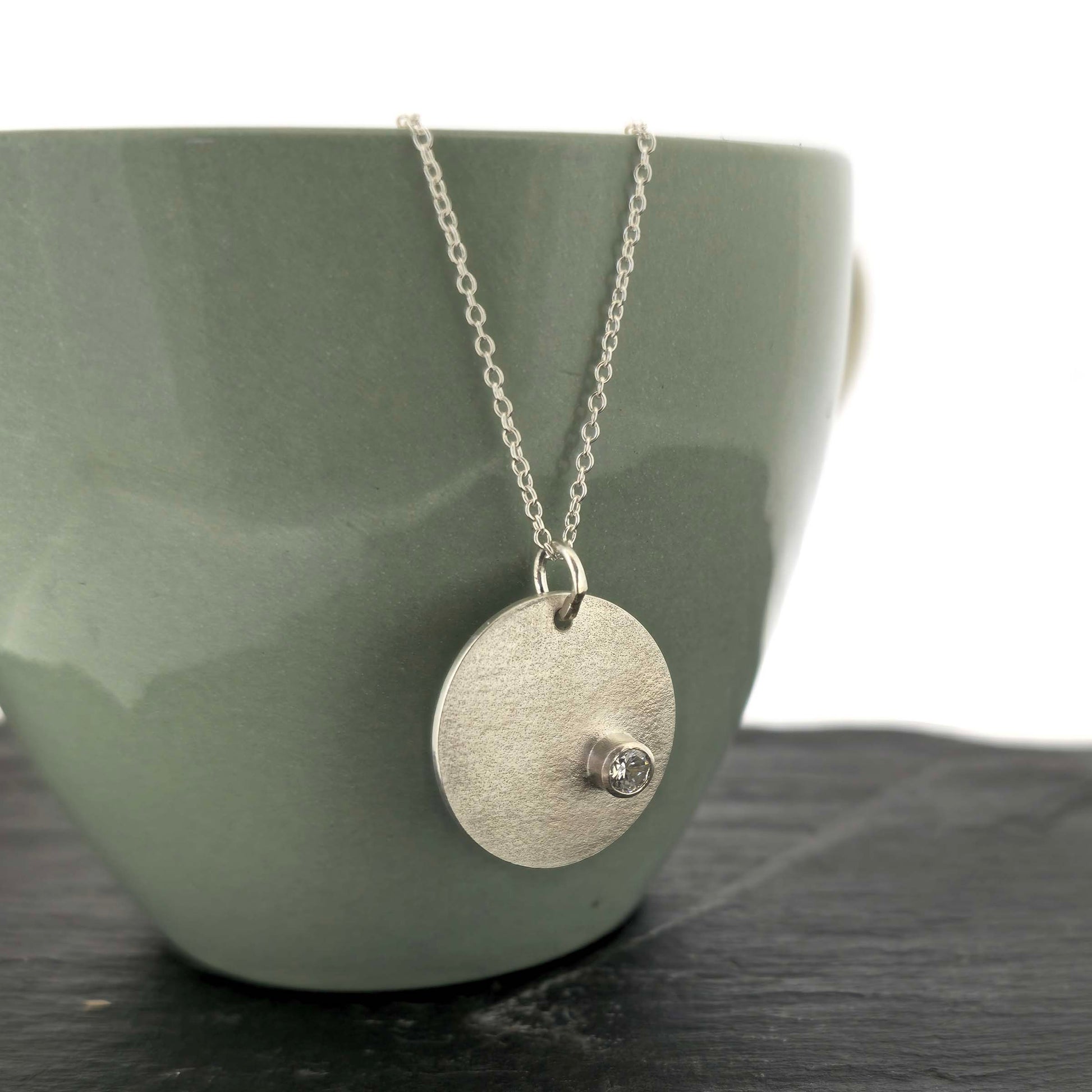 A silver round disc pendant with a matte finish and a sparkly clear CZ set off centre. On a silver chain. Pictured on a cup.