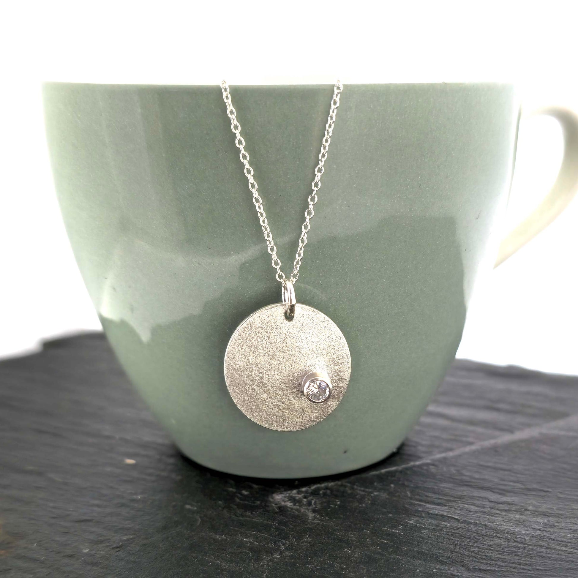 A silver round disc pendant with a matte finish and a sparkly clear CZ set off centre. On a silver chain. Pictured on a cup.