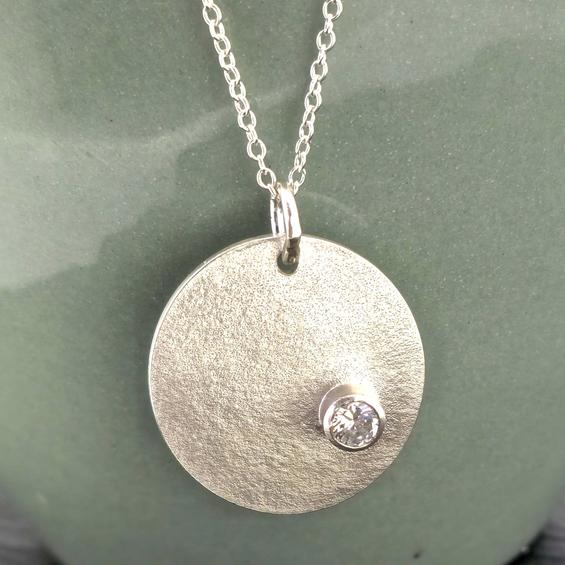 A silver round disc pendant with a matte finish and a sparkly clear CZ set off centre. On a silver chain. Front view.