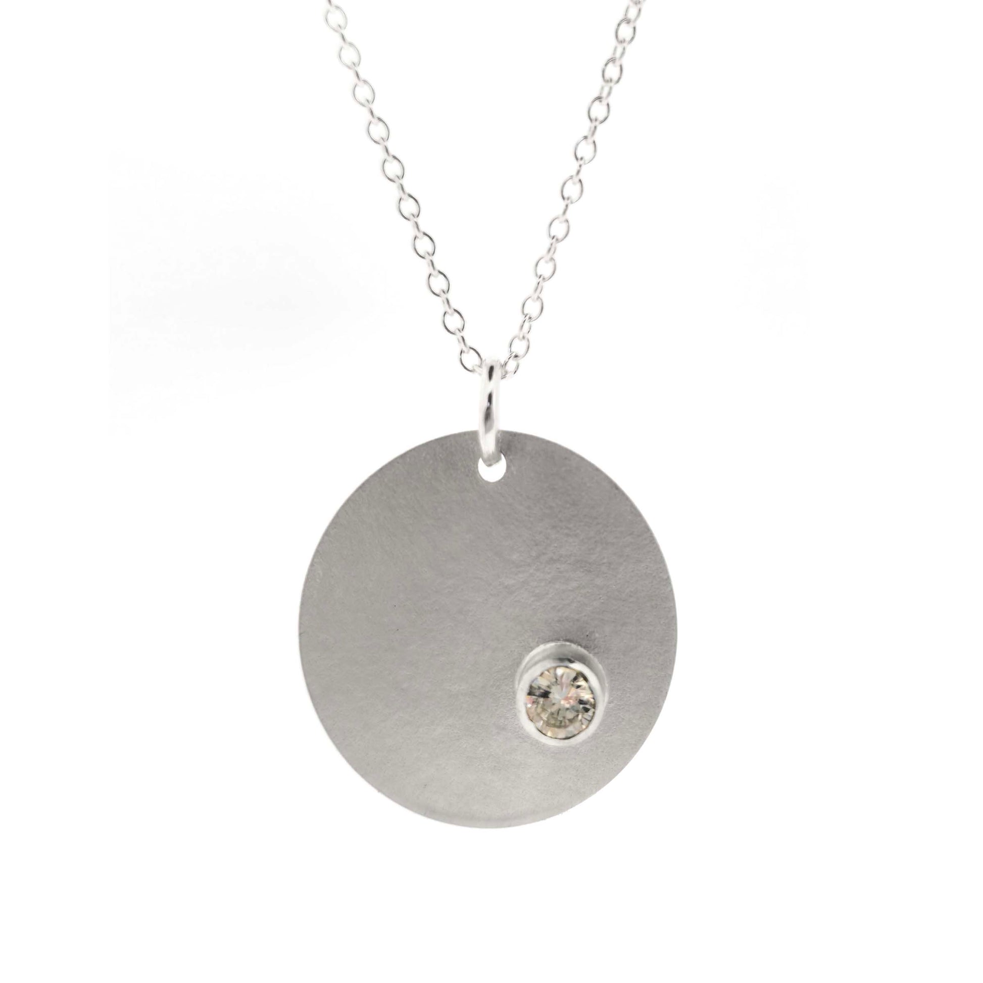 A silver round disc pendant with a matte finish and a sparkly clear CZ set off centre. On a silver chain. Front view.