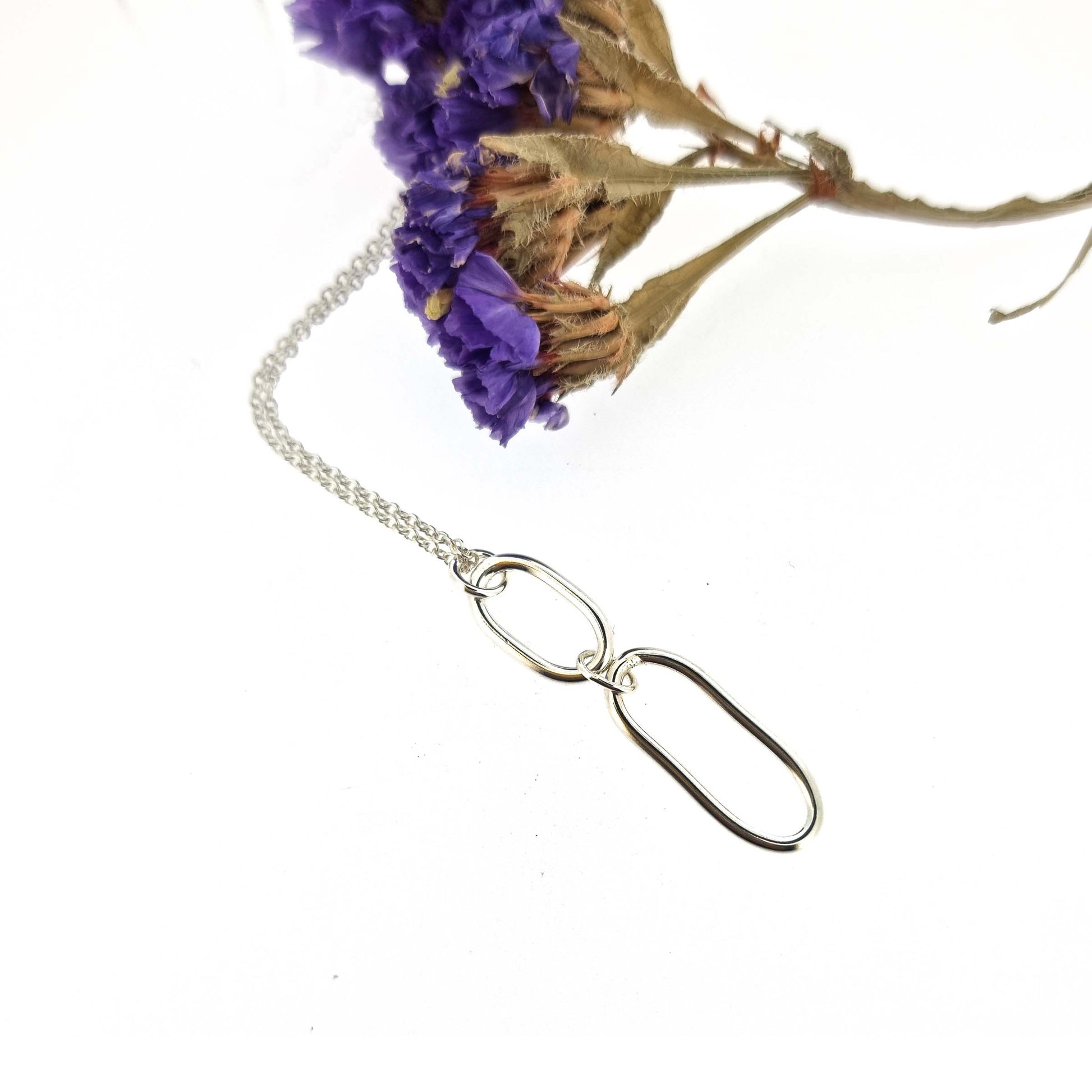 A silver pendant featuring 2 open oblong shapes on a silver chain. Pictured with flowers.