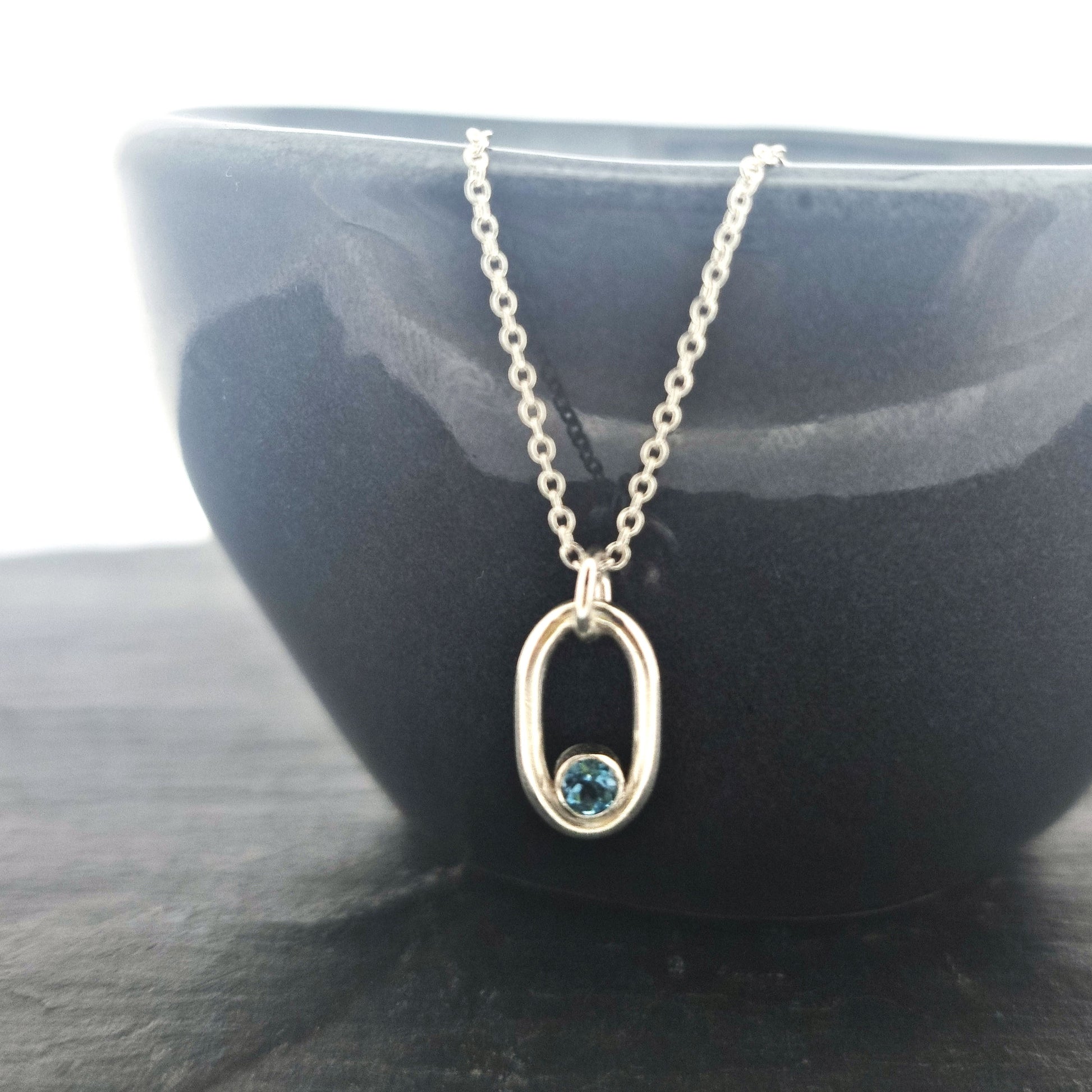 Silver open oblong pendant with a blue topaz gemstone set in the curve on a silver chain. Pictured on a black bowl.