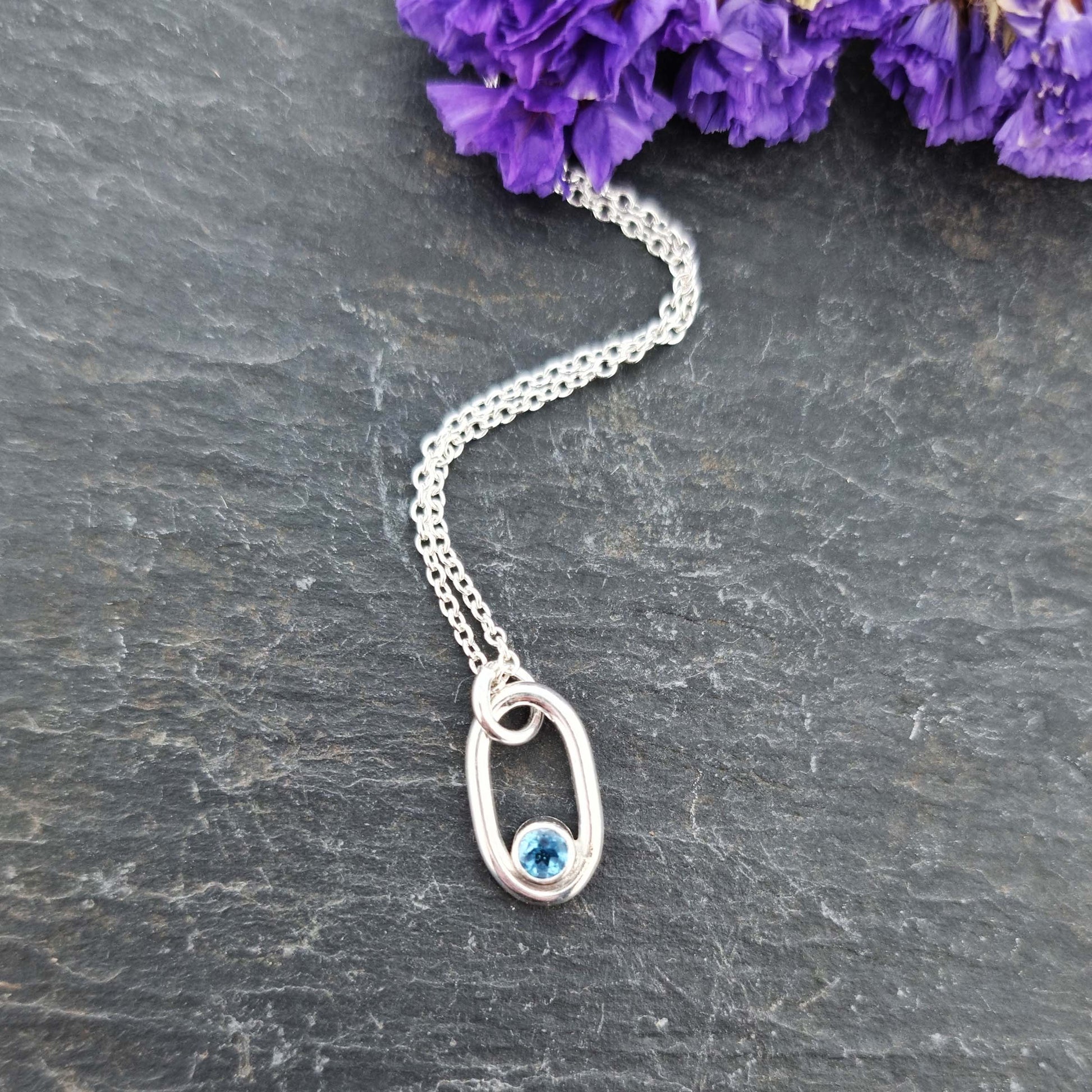 Silver open oblong pendant with a blue topaz gemstone set in the curve on a silver chain. Pictured with flowers.