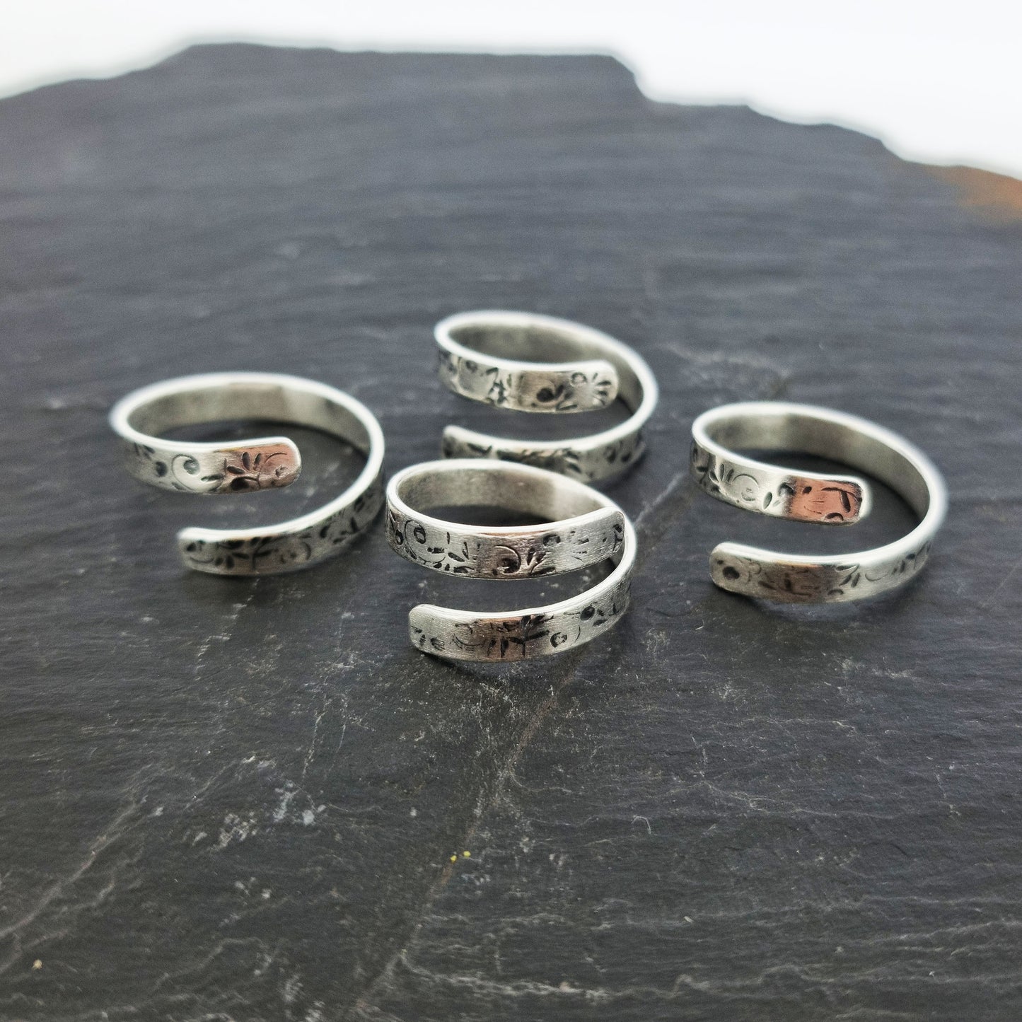 A group of flat silver wrap rings with a leaf, bud and stalk pattern. Shown on slate.