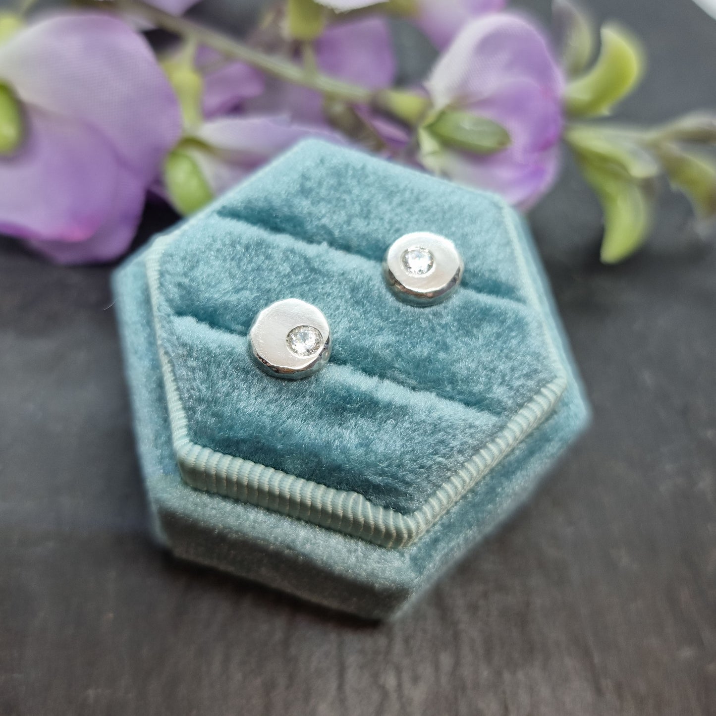 Silver flat 'pebble' stud earrings with a round CZ set off centre. In jewellery box with flowers.