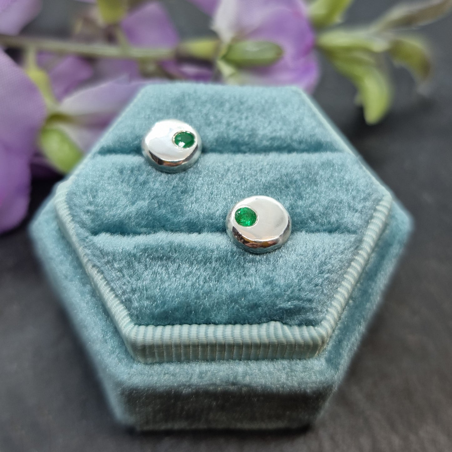 Round silver pebble stud earrings with green emerald gemstones set off-centre. Pictured in a jewellery box with flowers.