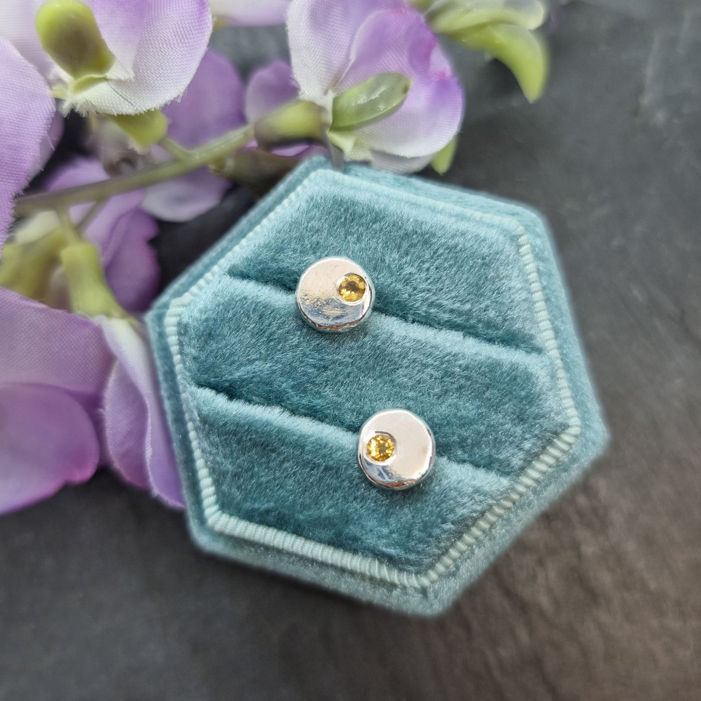 Round silver pebble stud earrings with yellow citrine gemstones flush set off-centre. Pictured in a jewellery box with flowers.