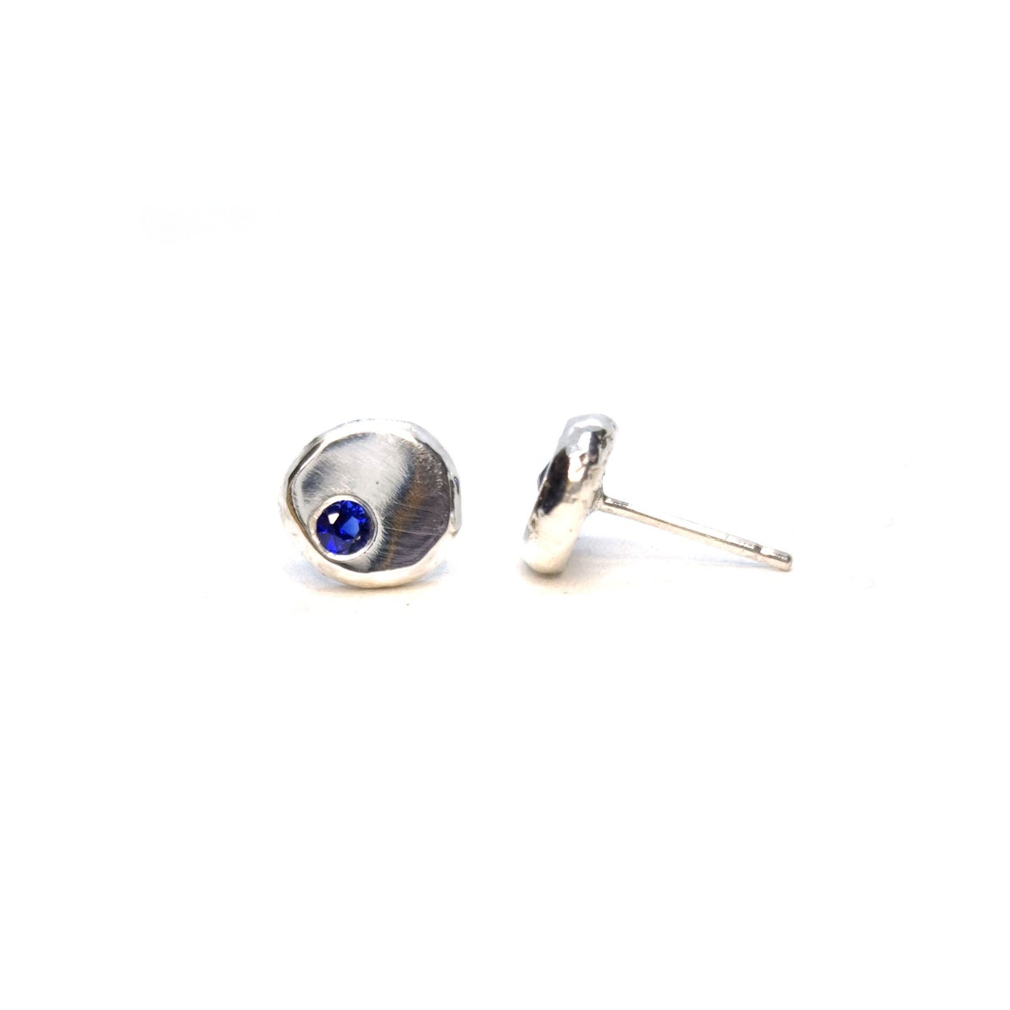 Round pebble stud earrings set with dark blue sapphire gemstones off -centre.