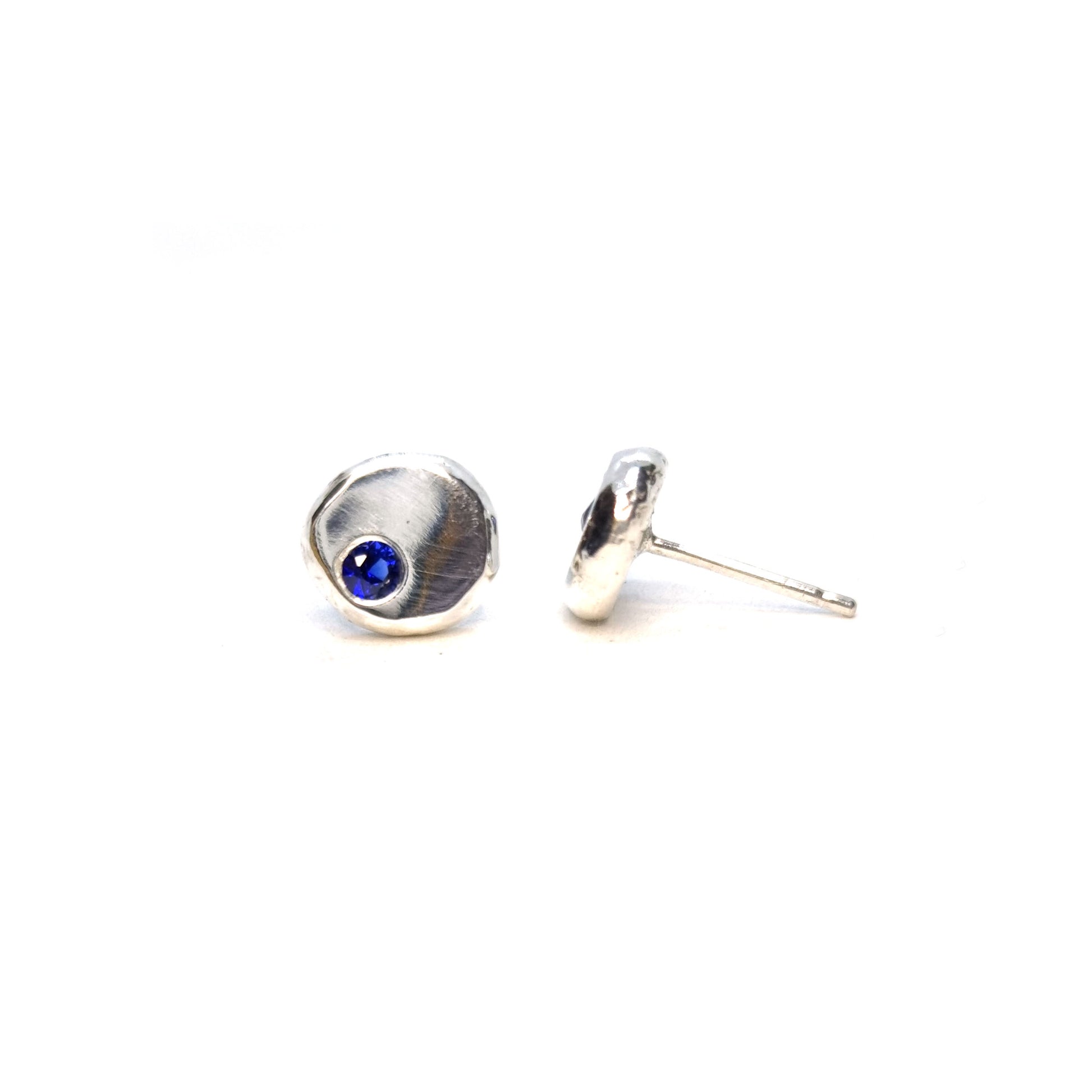 Round pebble stud earrings set with dark blue sapphire gemstones off -centre.