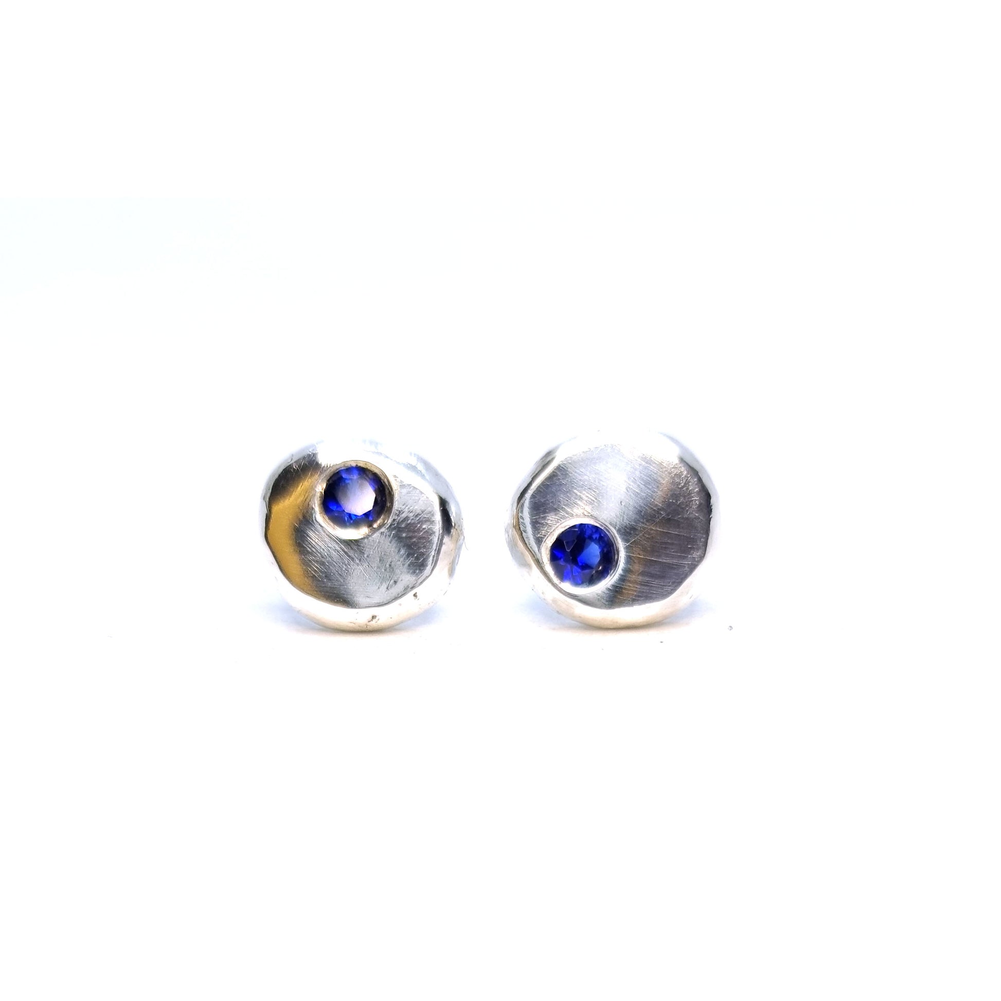 Round pebble stud earrings set with dark blue sapphire gemstones off -centre.
