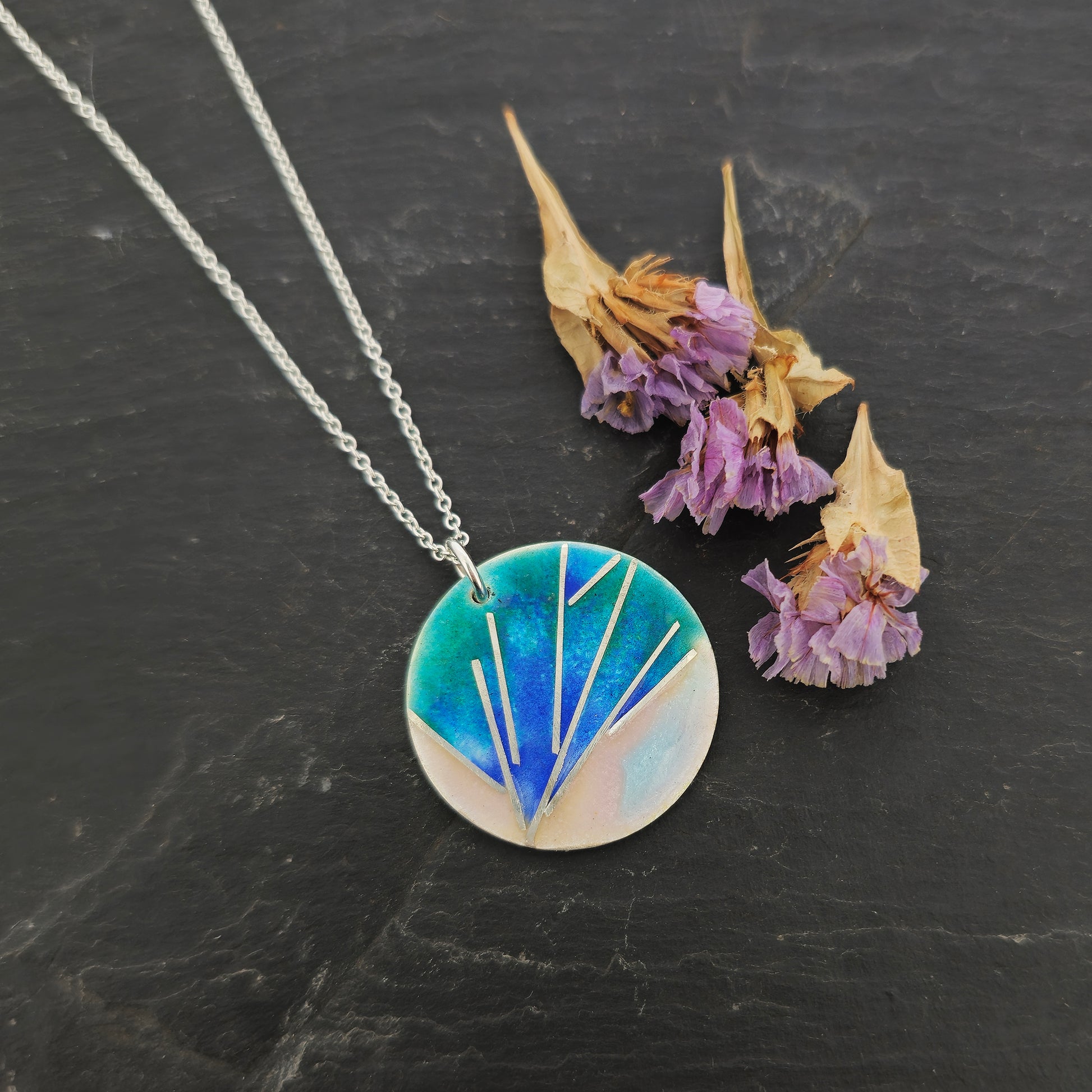 A round silver pendant with a spray of silver lines and blue, pink, and green enamel. On a silver chain. Pictured on slate with flowers.