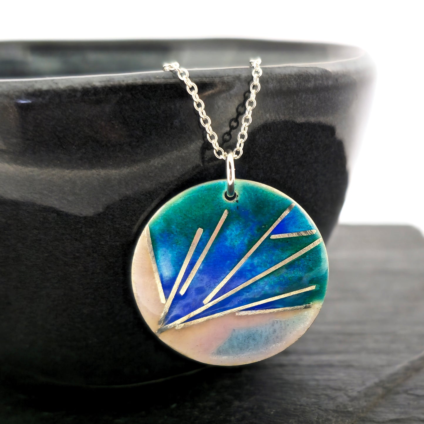 A round silver pendant with a spray of silver lines and blue, pink, and green enamel. On a silver chain. Pictured on a bowl.