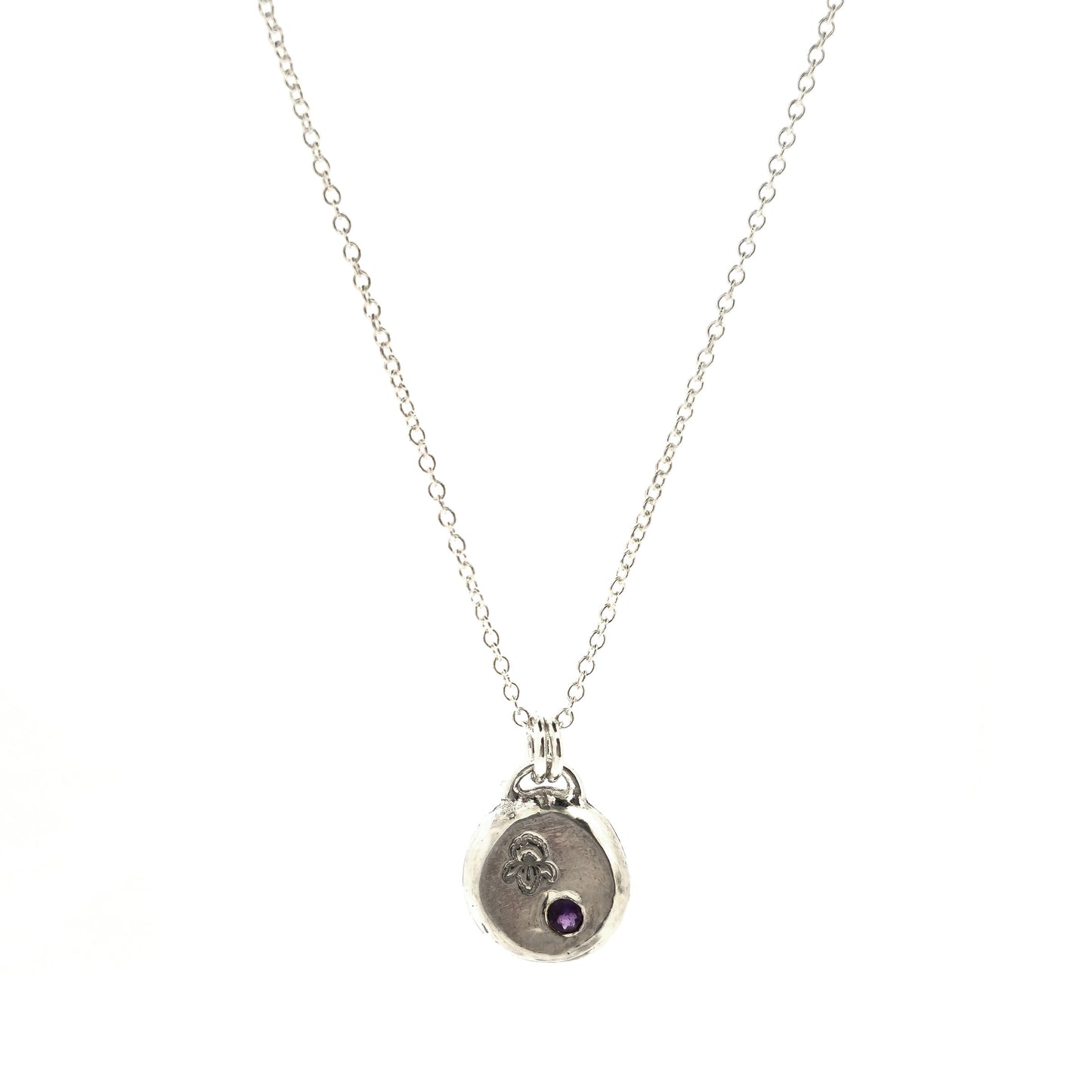 A handmade round silver pendant with a violet flower (February birth flower) engraved on it and a round purple amethyst (February birthstone) flush set into it. On a silver chain. On a white background. Mijoux Jewellery.