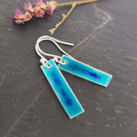 Rectangular silver drop earrings with a turquoise enamel background and a speckled line of dark blue enamel in the middle. On silver ear hooks. Shown on slate with flowers.