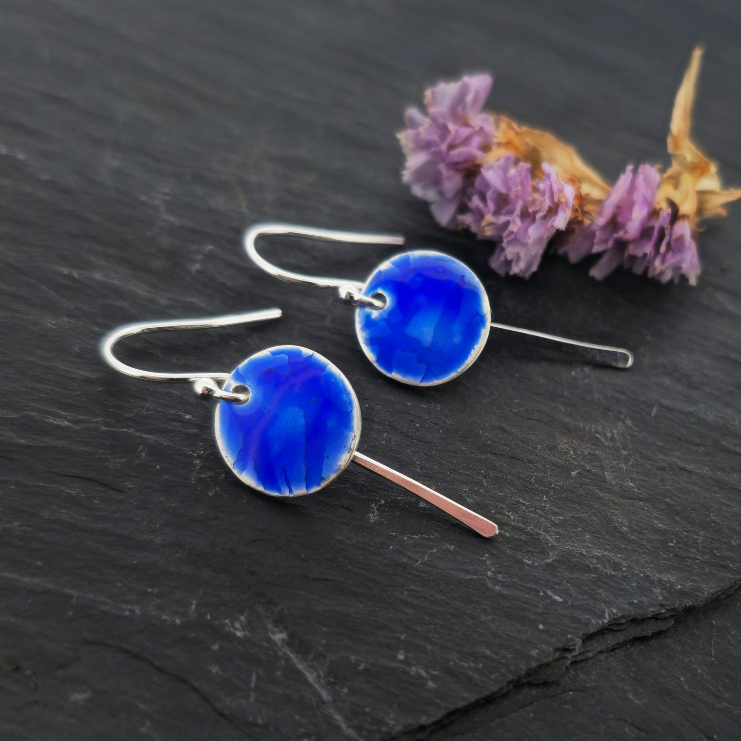 Silver drop earrings featuring a rustic round disc with a lined pattern covered with dark blue enamel. slim silver tag hangs behind and below. On silver ear hooks. Shown on slate with flowers.