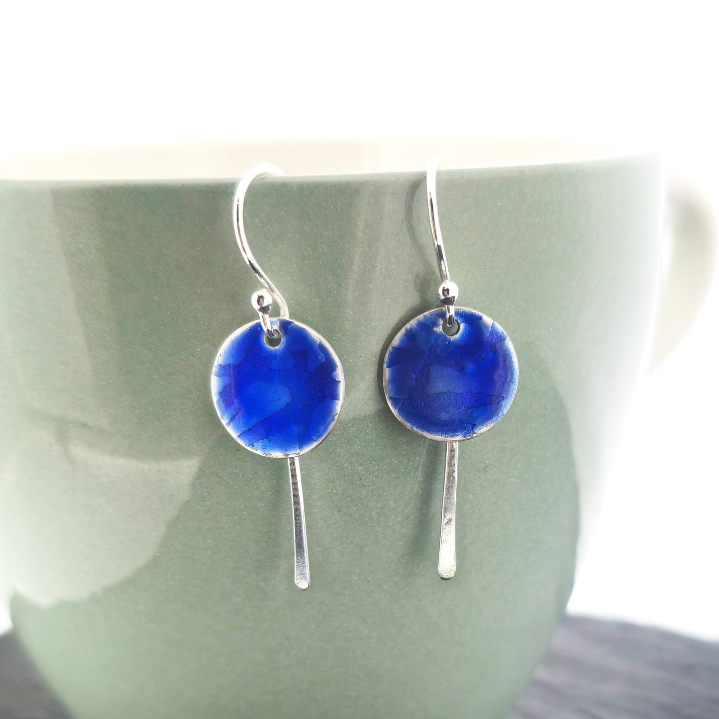 Silver drop earrings featuring a rustic round disc with a lined pattern covered with dark blue enamel. slim silver tag hangs behind and below. On silver ear hooks. Shown on a light green cup.