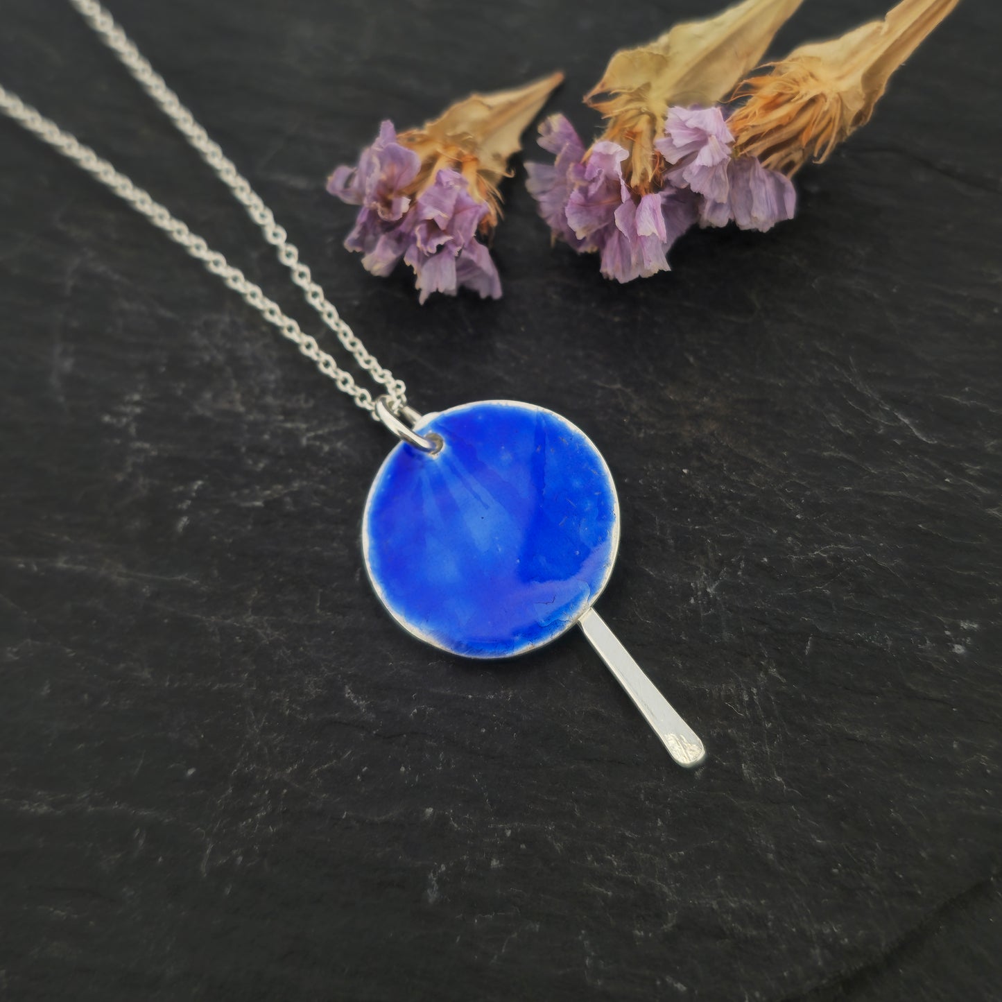 A silver pendant with a round disc that has a lined pattern covered in royal blue enamel. A silver long strip is suspended behind and comes own below the disc. On a silver chain. Shown on slate with flowers.