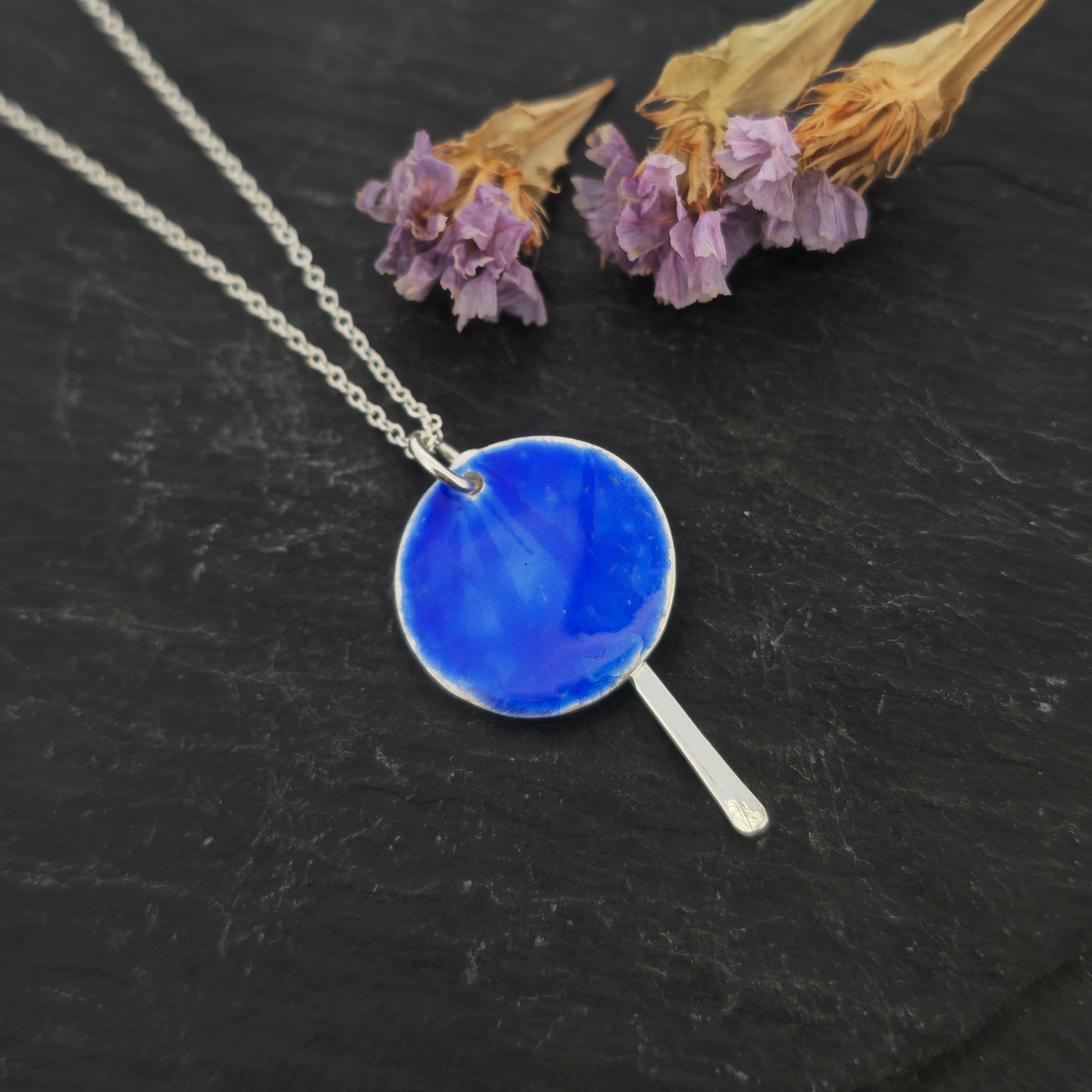 A silver pendant with a round disc that has a lined pattern covered in royal blue enamel. A silver long strip is suspended behind and comes own below the disc. On a silver chain. Shown on slate with flowers.