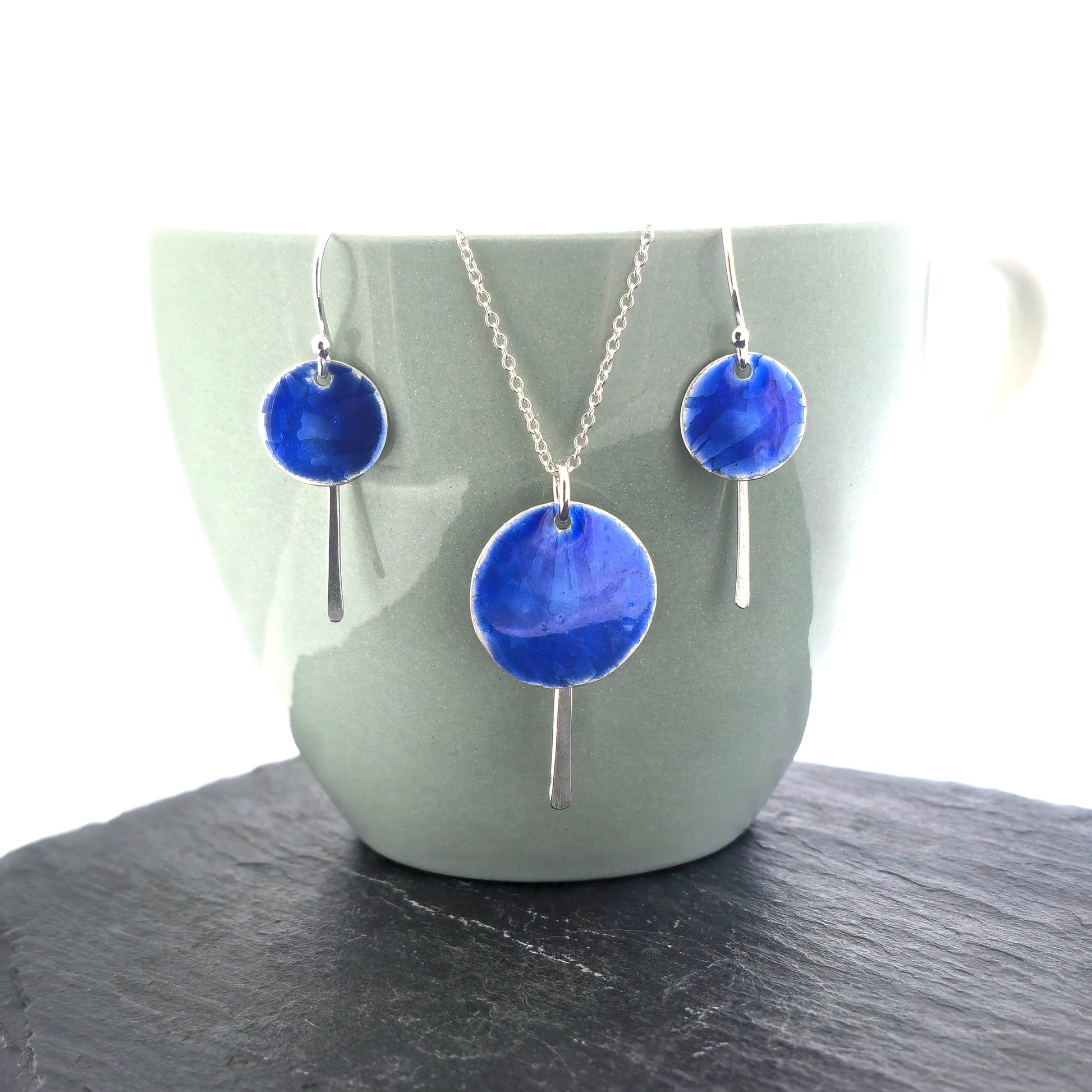 A silver pendant with a round disc that has a lined pattern covered in royal blue enamel. A silver long strip is suspended behind and comes own below the disc. On a silver chain. Shown on a cup with matching drop earrings.