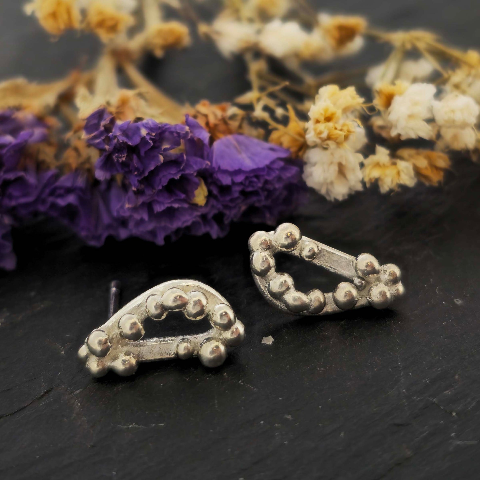Silver asymmetric stud earrings with an open soft triangle shape and silver balls on a black background with dried flowers.