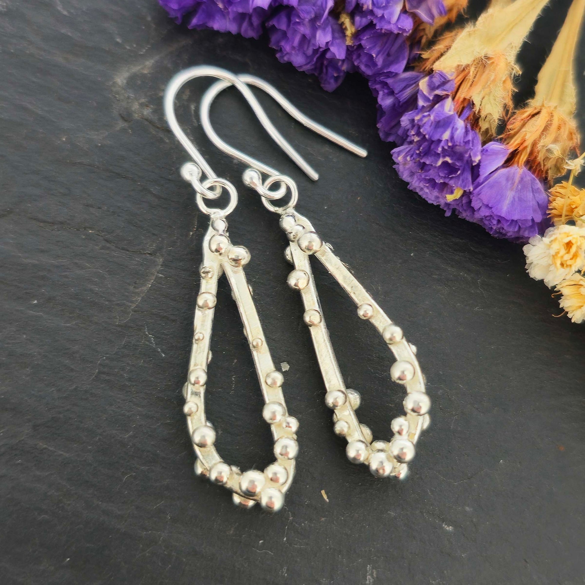 Silver drop earrings in a asymmetrical open teardrop shape with silver balls. Shown on a black background with dried flowers.