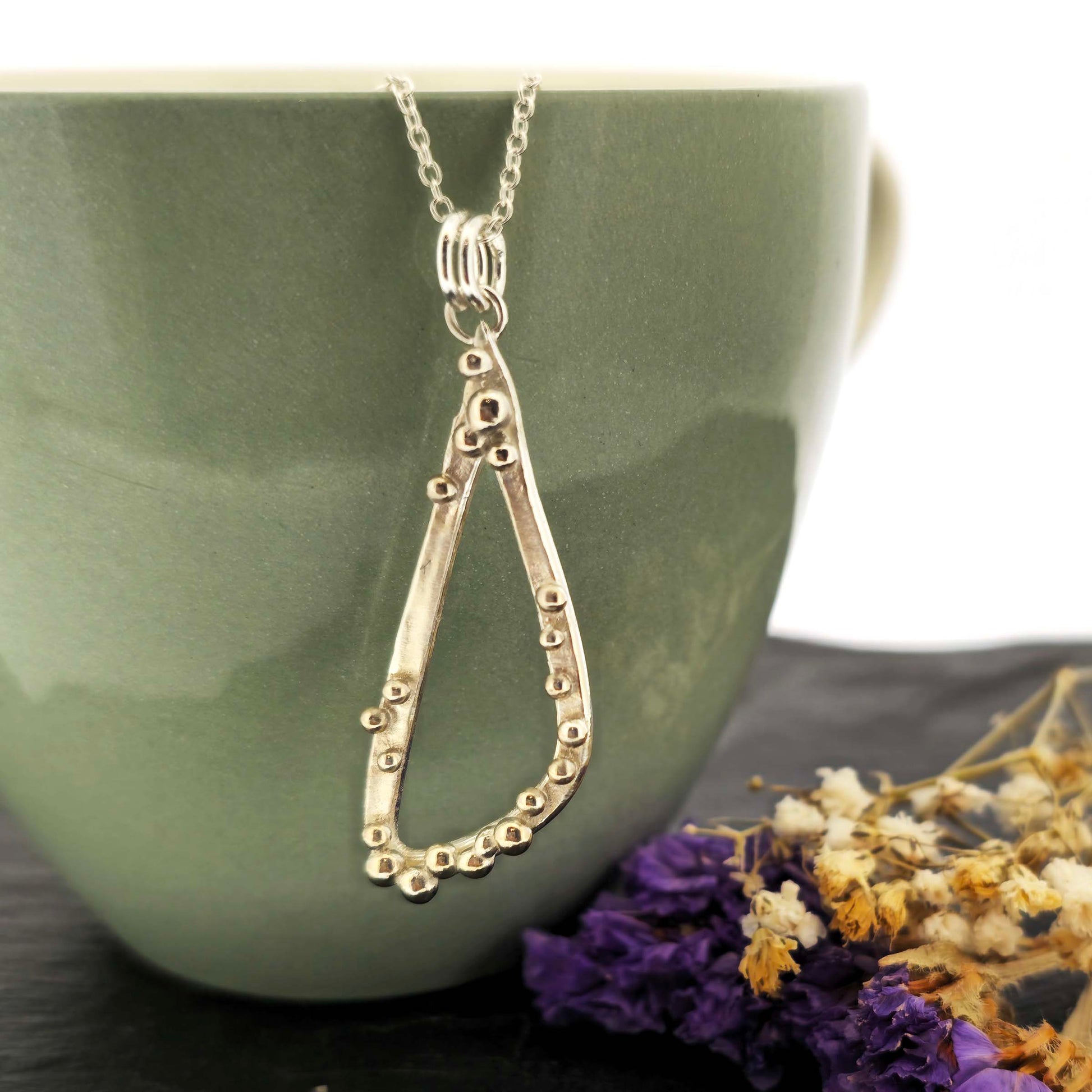 A silver asymmetrical pendant with a soft open triangle shape and silver balls, suspended from a silver chain. Shown on a green bowl with dried flowers.